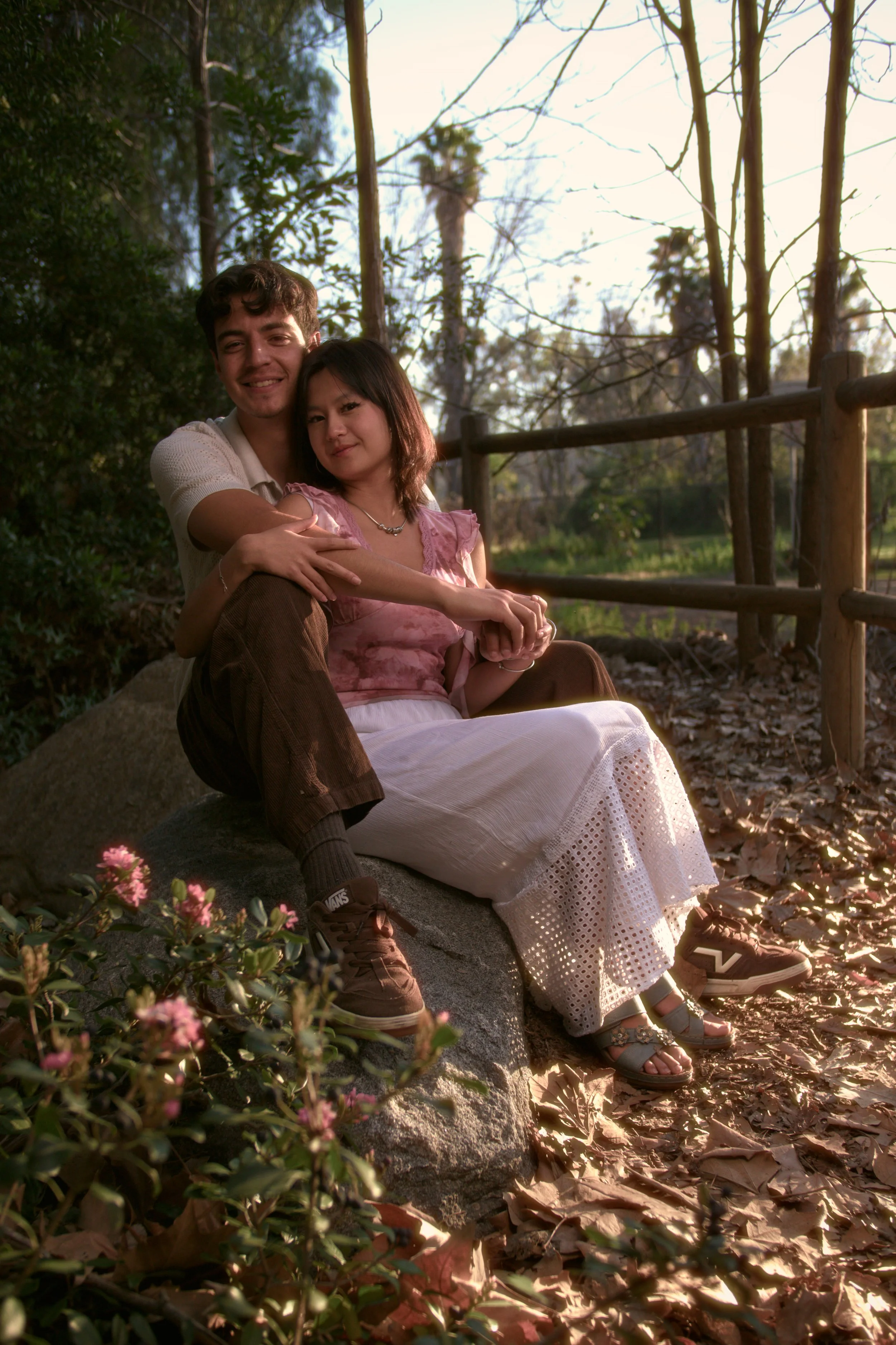 A young man and woman sitting on a large rock in a park, surrounded by trees and fallen leaves, sharing a moment during sunset.