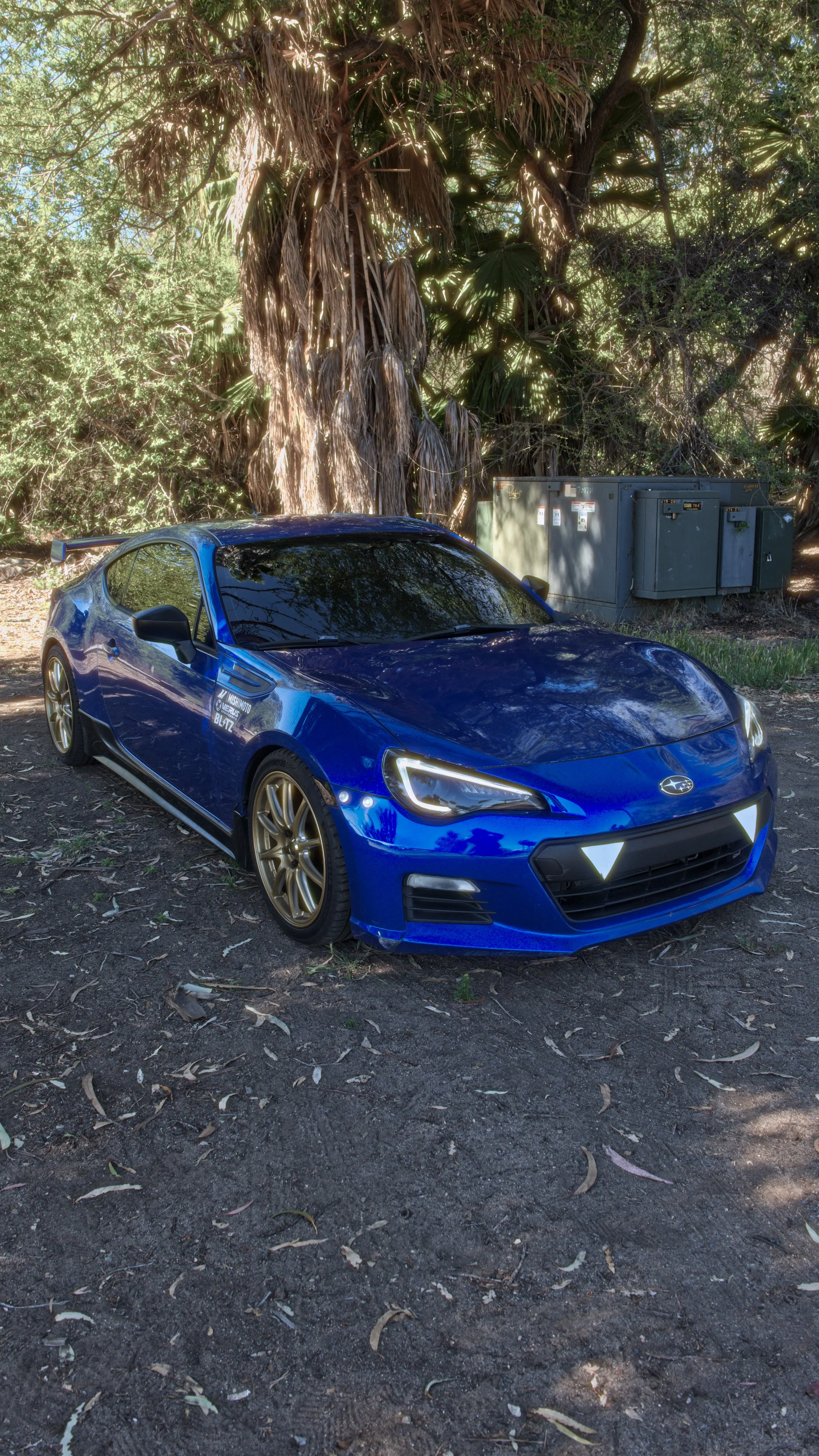 A blue Subaru sports car with gold wheels parked on dirt ground under trees, with a large tree and utility boxes in the background.