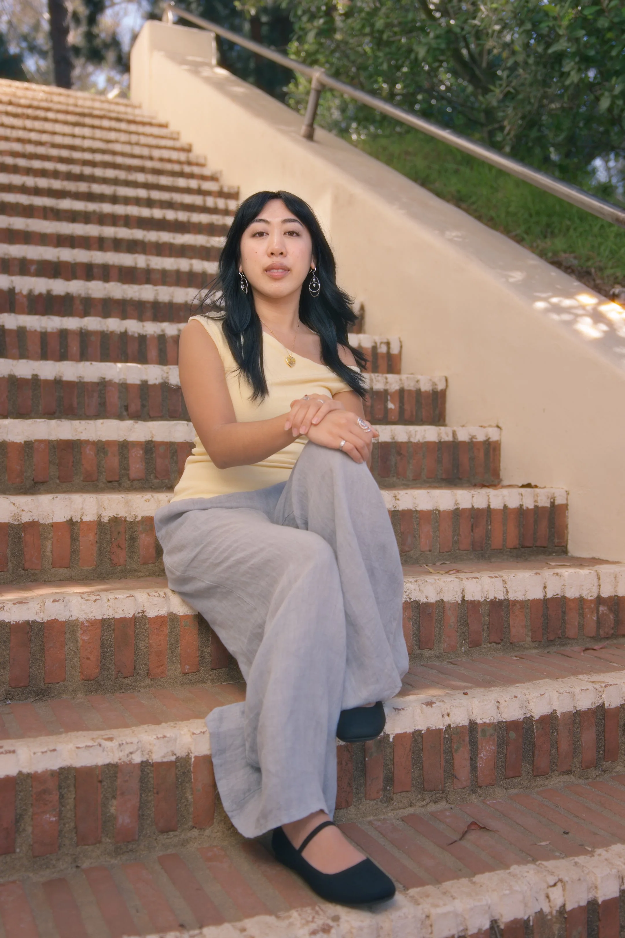 A young woman with black hair and tan skin sitting on brick stairs outdoors, wearing a yellow sleeveless top, gray wide-leg pants, black shoes, and silver jewelry, with trees and a cream-colored wall in the background.