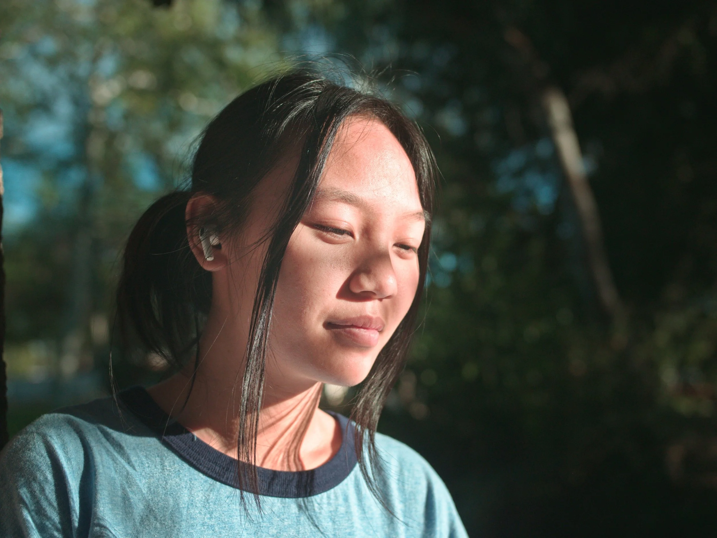 Close-up of a young woman with black hair wearing a blue T-shirt and wireless earbuds, looking down with sunlight illuminating her face, in an outdoor setting with blurred trees in the background.