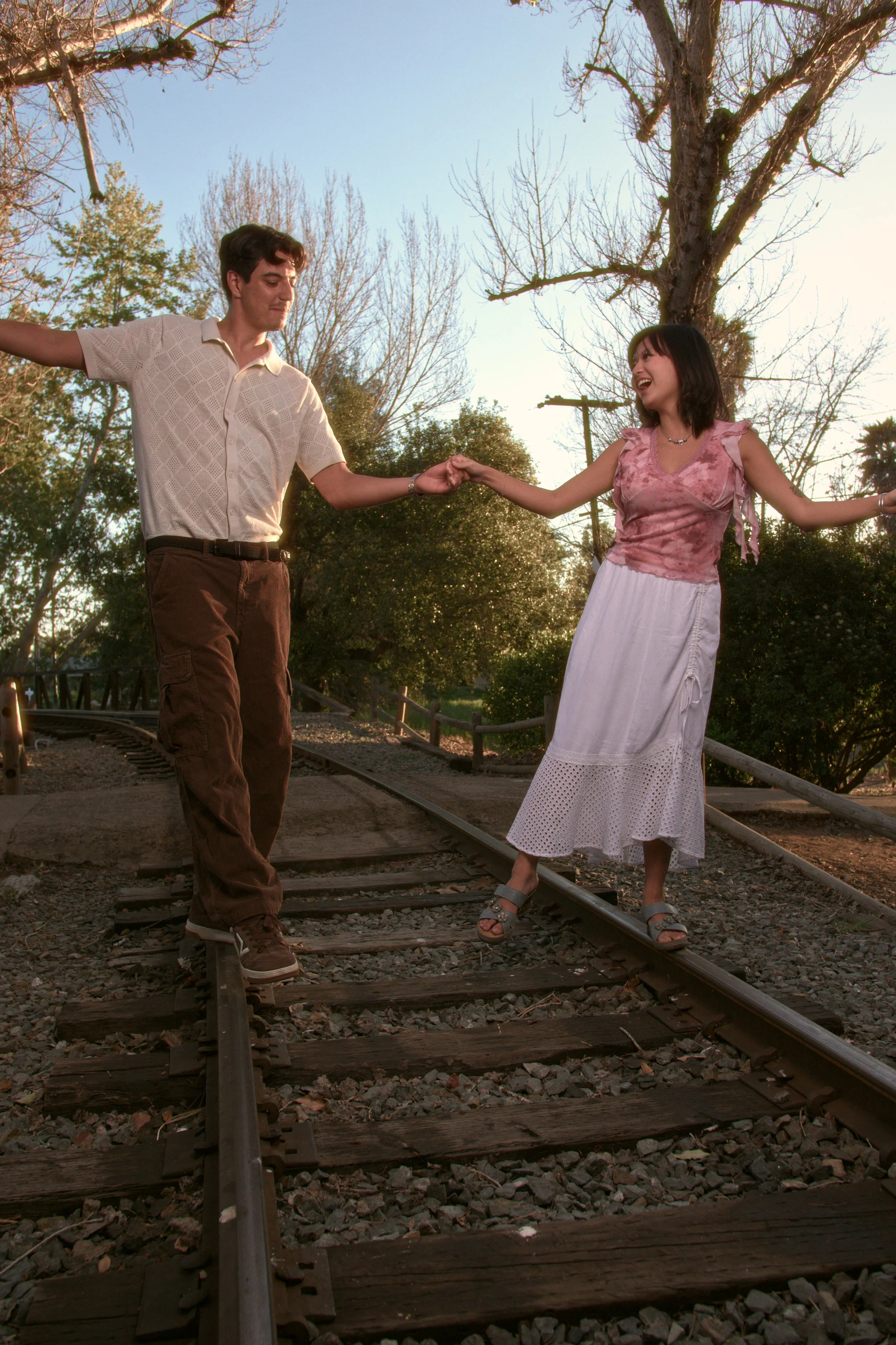 A young man and a young woman are balancing and holding hands while walking on railroad tracks outdoors, with trees and a clear sky in the background.