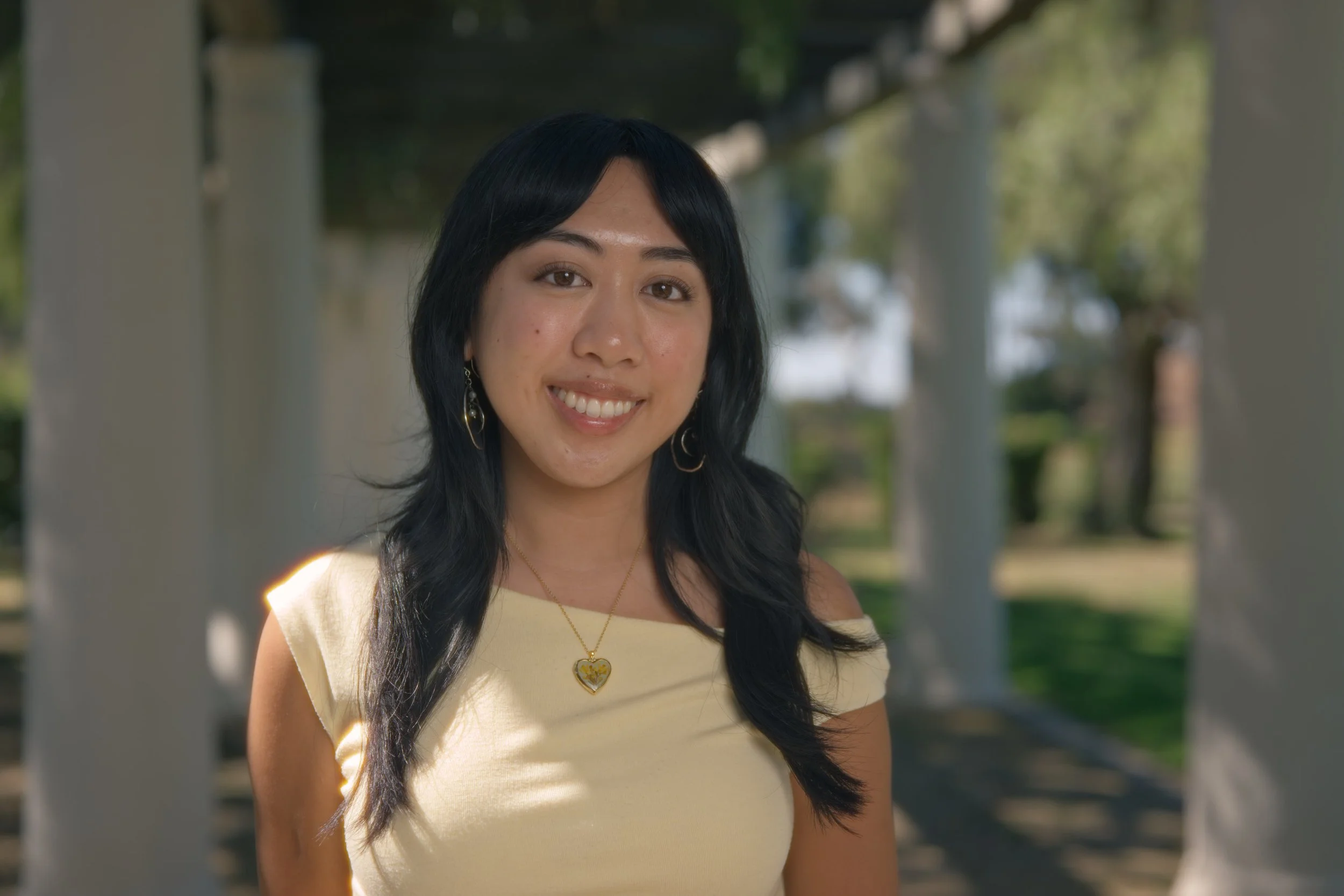 A woman with black hair and earrings, smiling outdoors, wearing a yellow top and a necklace with a heart pendant.