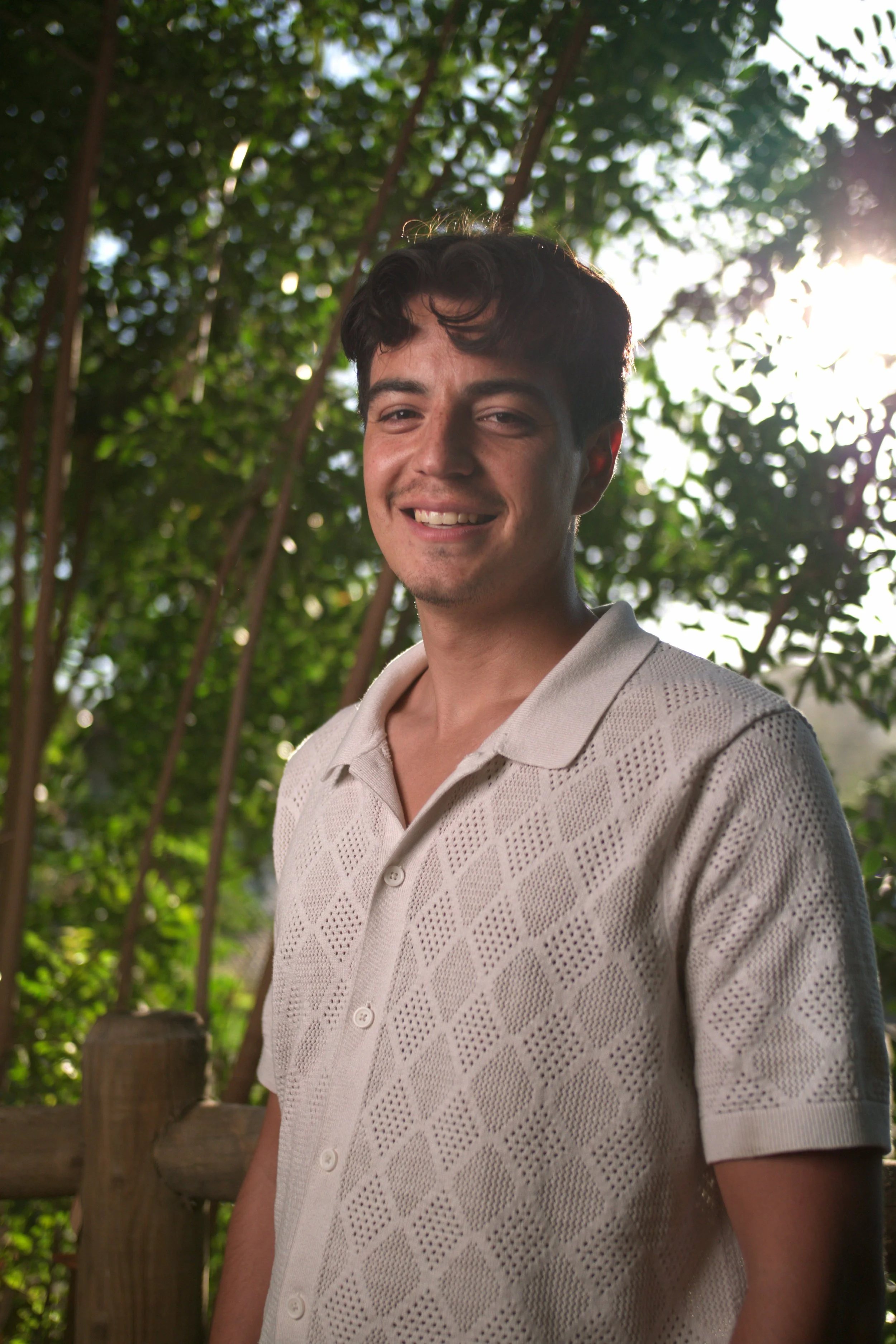 A young man with dark hair, smiling, standing outdoors near greenery and sunlight.