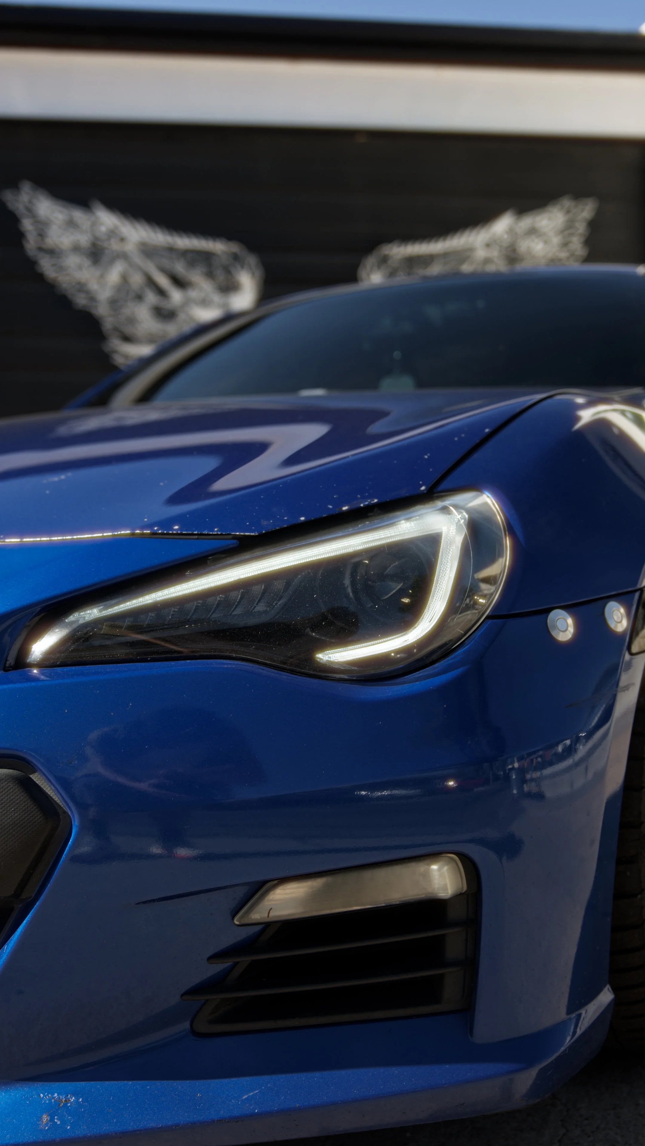 Close-up of the front of a blue sports car, showing the headlight, front grille, and part of the hood, with a black wall in the background featuring a winged skull decal.