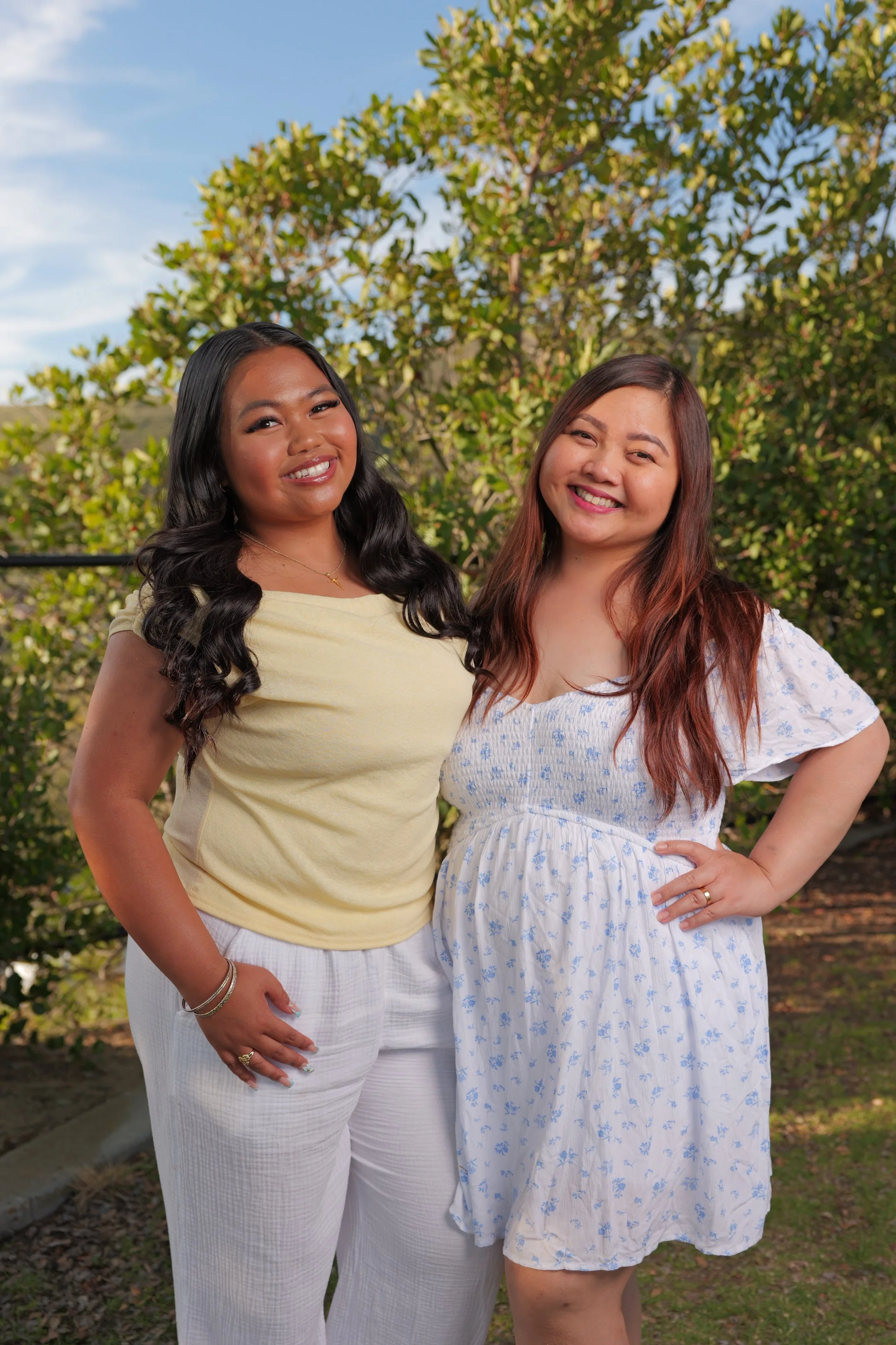 Two smiling women standing outdoors in front of green trees, one with long black hair in a yellow top and white pants, the other with long reddish-brown hair in a white dress with blue flowers.