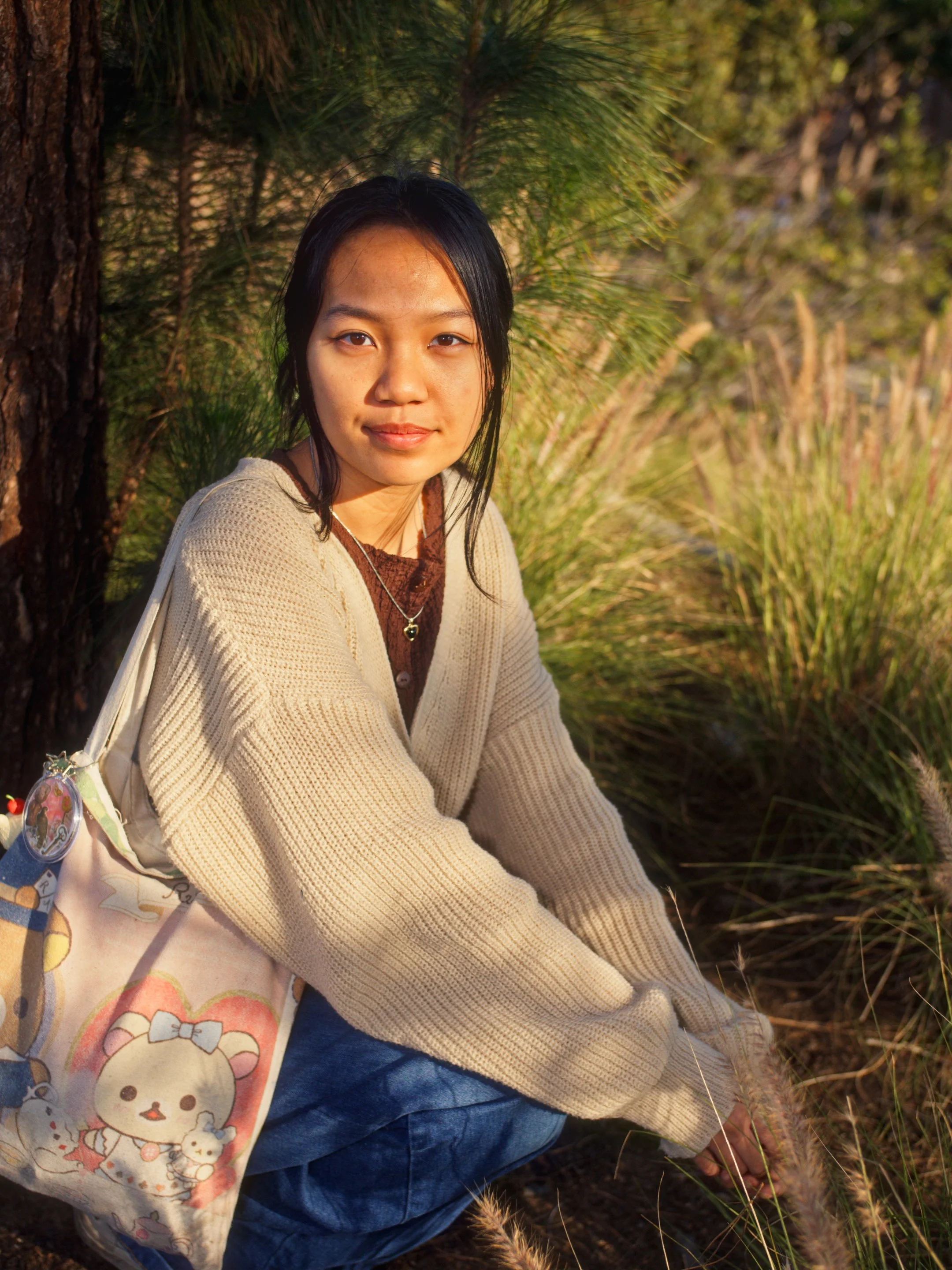 A young woman with long black hair, wearing a beige sweater over a brown top, sitting outdoors amidst greenery and tall grass, carrying a colorful tote bag with cartoon characters.