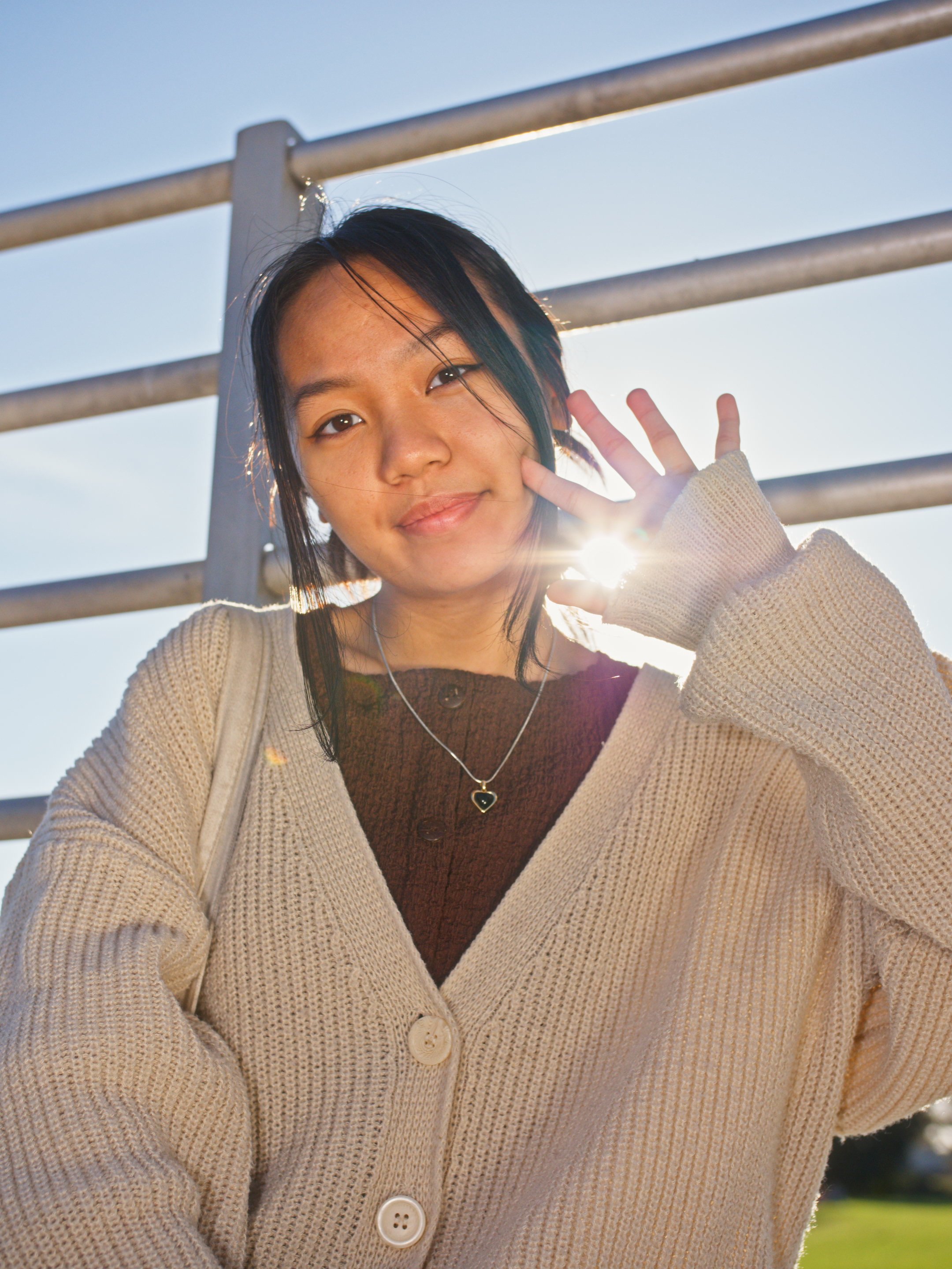 A young woman with black hair and a light complexion smiling at the camera, giving a peace sign with her right hand, outdoors with metal railing and bright sunlight in the background.
