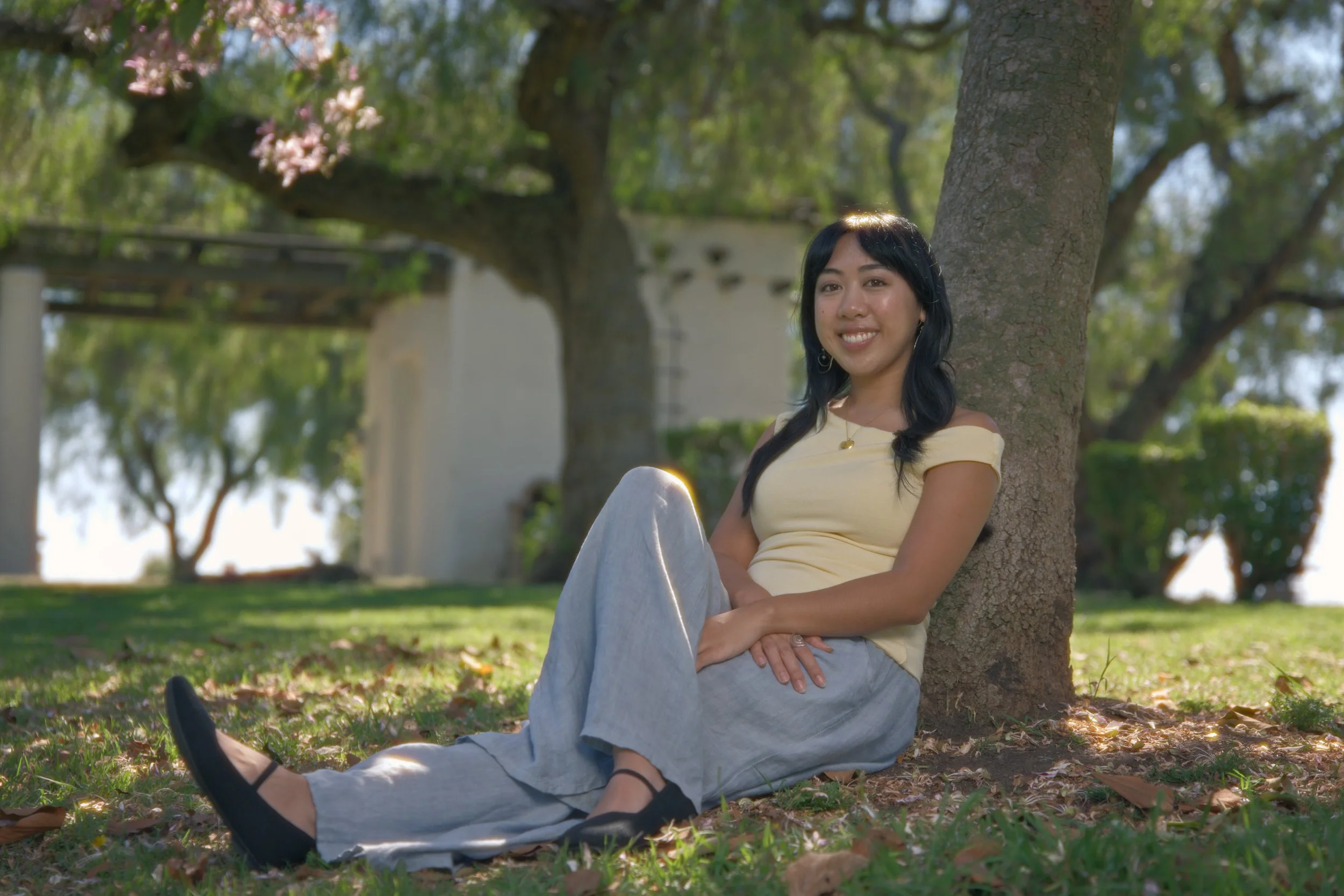 A young woman with dark hair smiling and sitting on grass against a tree in a park during daytime, wearing a yellow top and light gray pants.