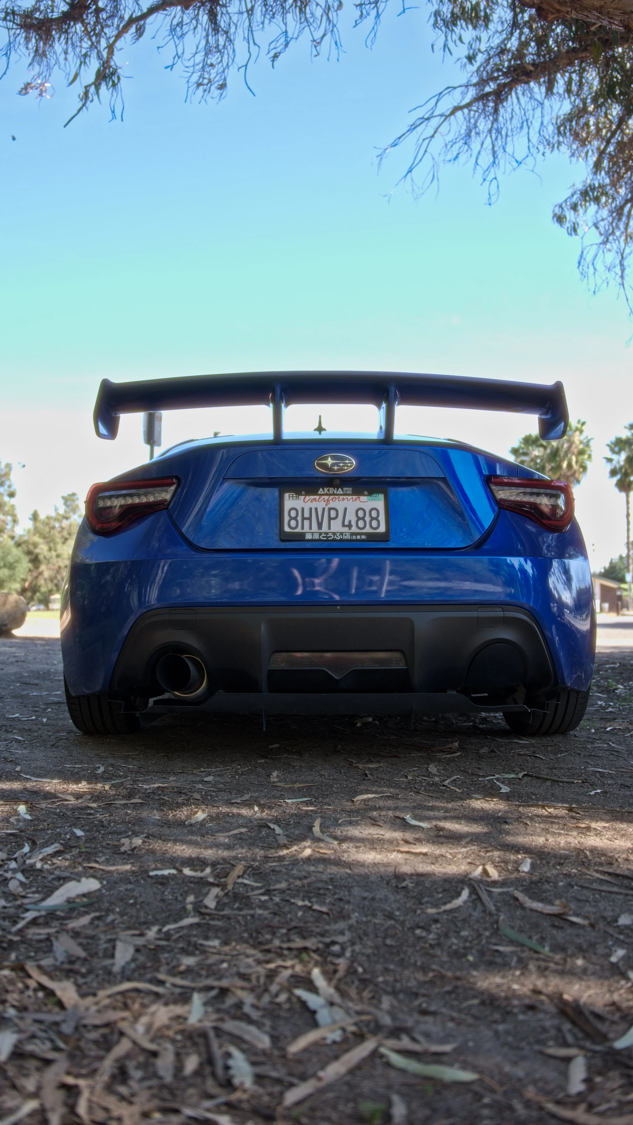 Rear view of a blue Subaru sports car with a large rear spoiler, parked on dirt ground with fallen leaves, under a partly cloudy sky with tree branches overhead.