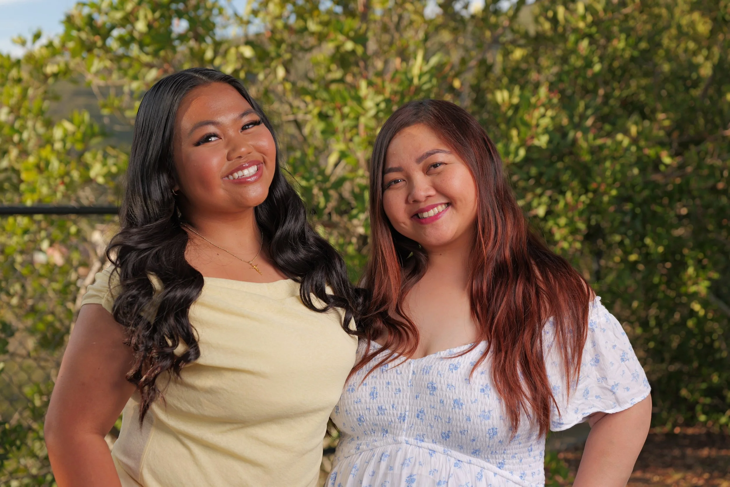Two women smiling outdoors with green trees in the background, one with long black hair and the other with long reddish-brown hair, both wearing casual summer dresses.