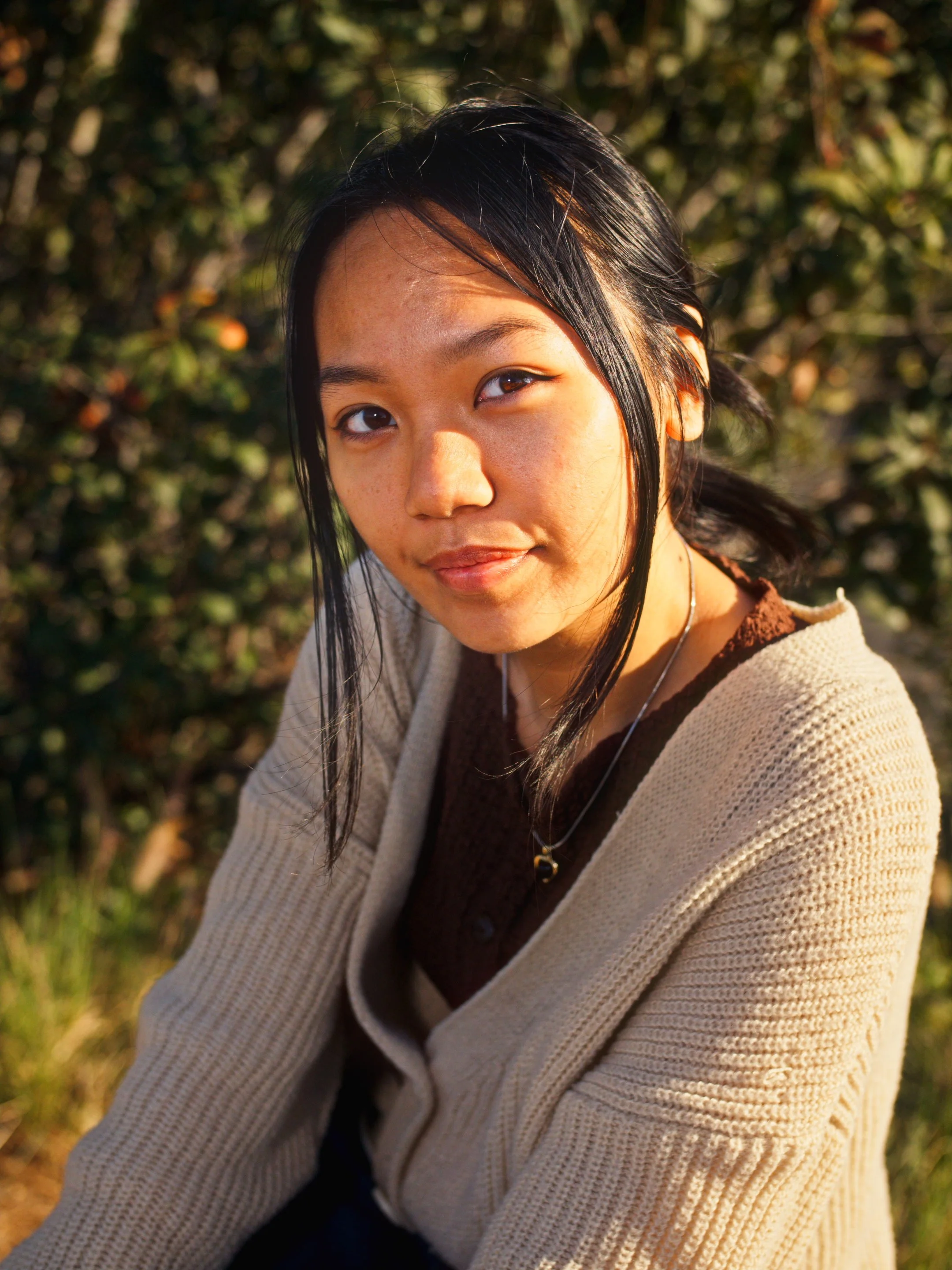 A young woman with black hair and light brown skin wearing a beige sweater and a necklace, sitting outdoors in sunlight with greenery in the background.