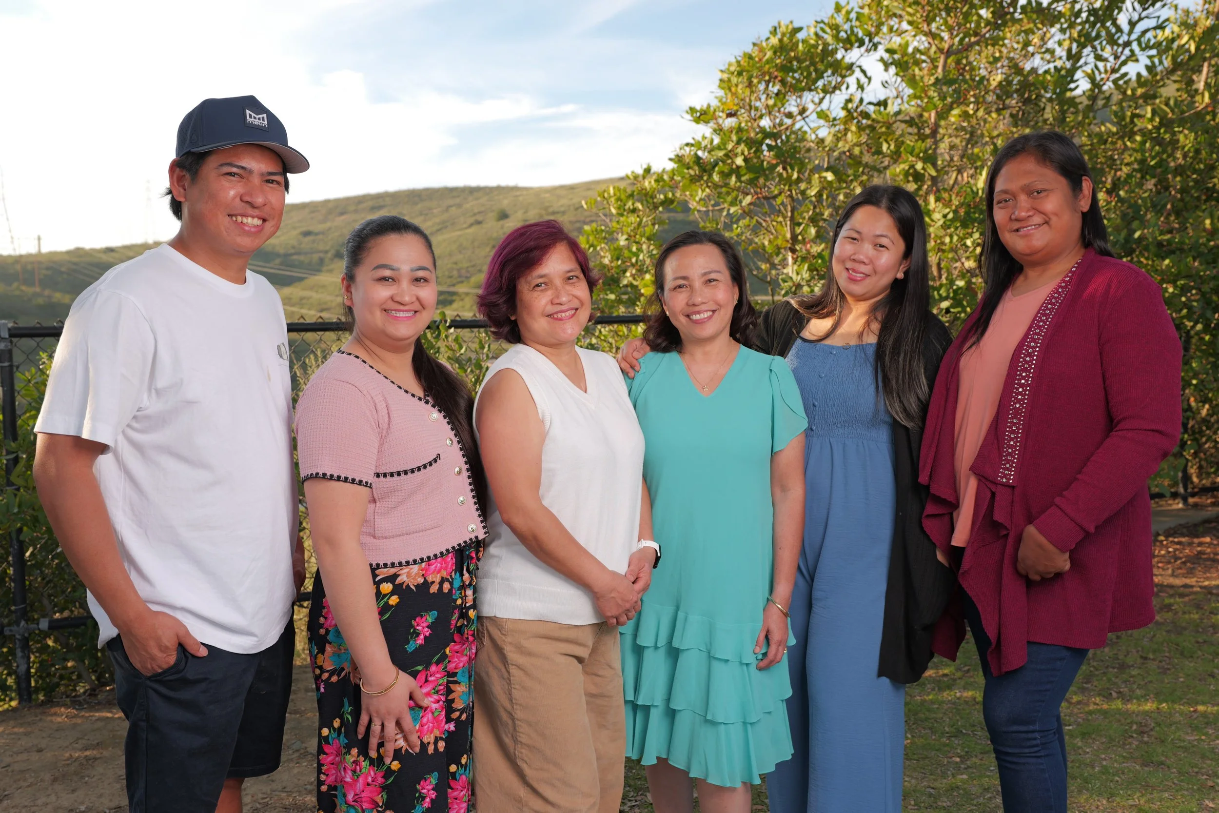 Seven people standing outdoors in front of a black fence and green trees with hills in the background. They are smiling and wearing casual clothing.