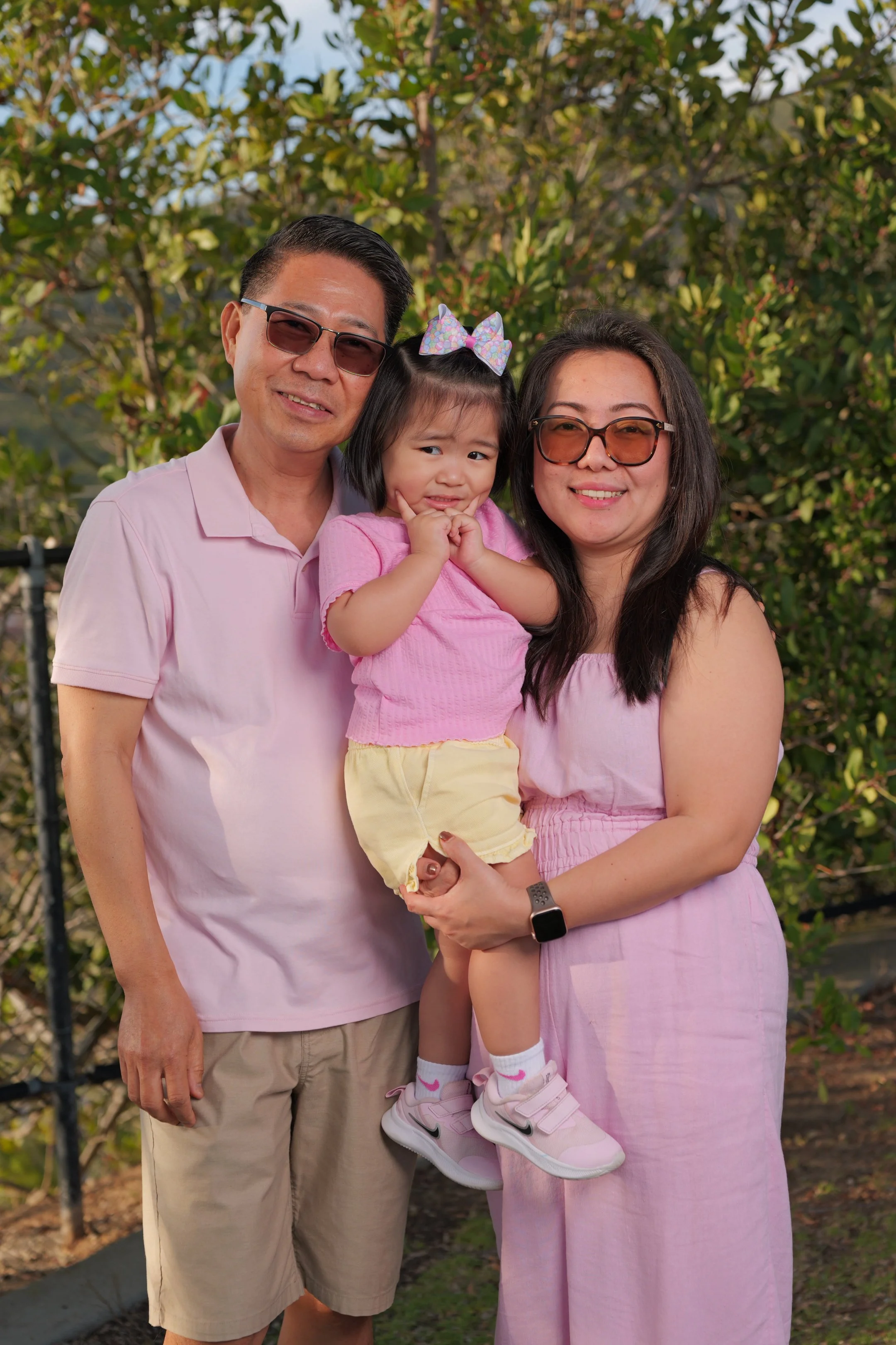 Family of three outdoors in a park, adults wearing pink and a young girl in pink and yellow, all smiling at the camera.