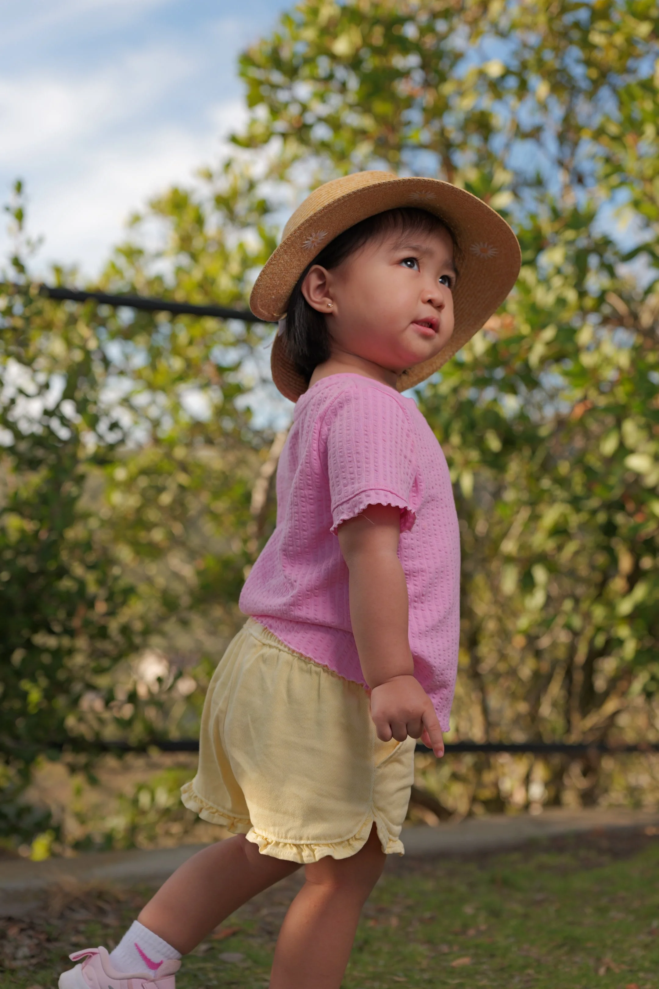 A young girl wearing a wide-brimmed straw hat, pink shirt, yellow shorts, white socks, and pink shoes, standing outdoors among trees with sunlight shining through the leaves.