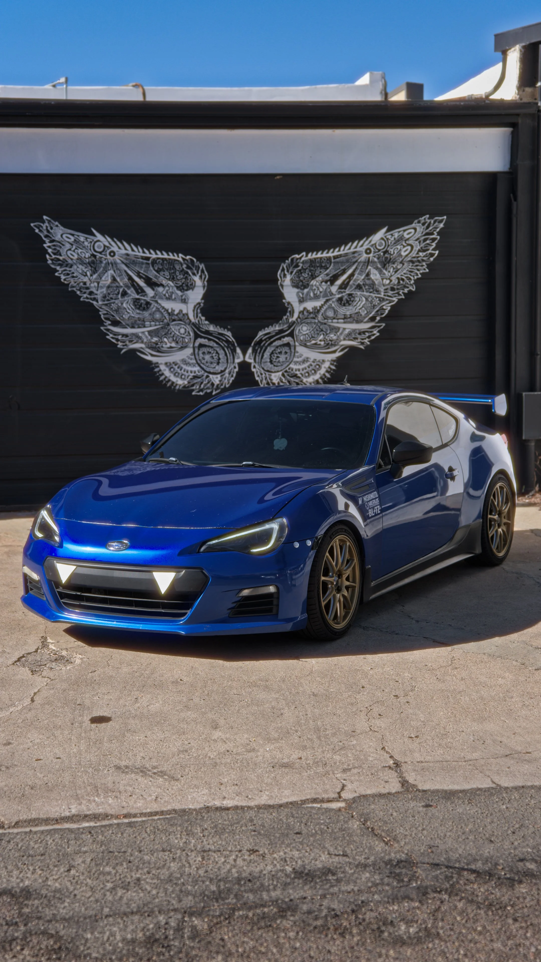 A blue sports car parked in front of a black wall with white winged butterfly artwork.