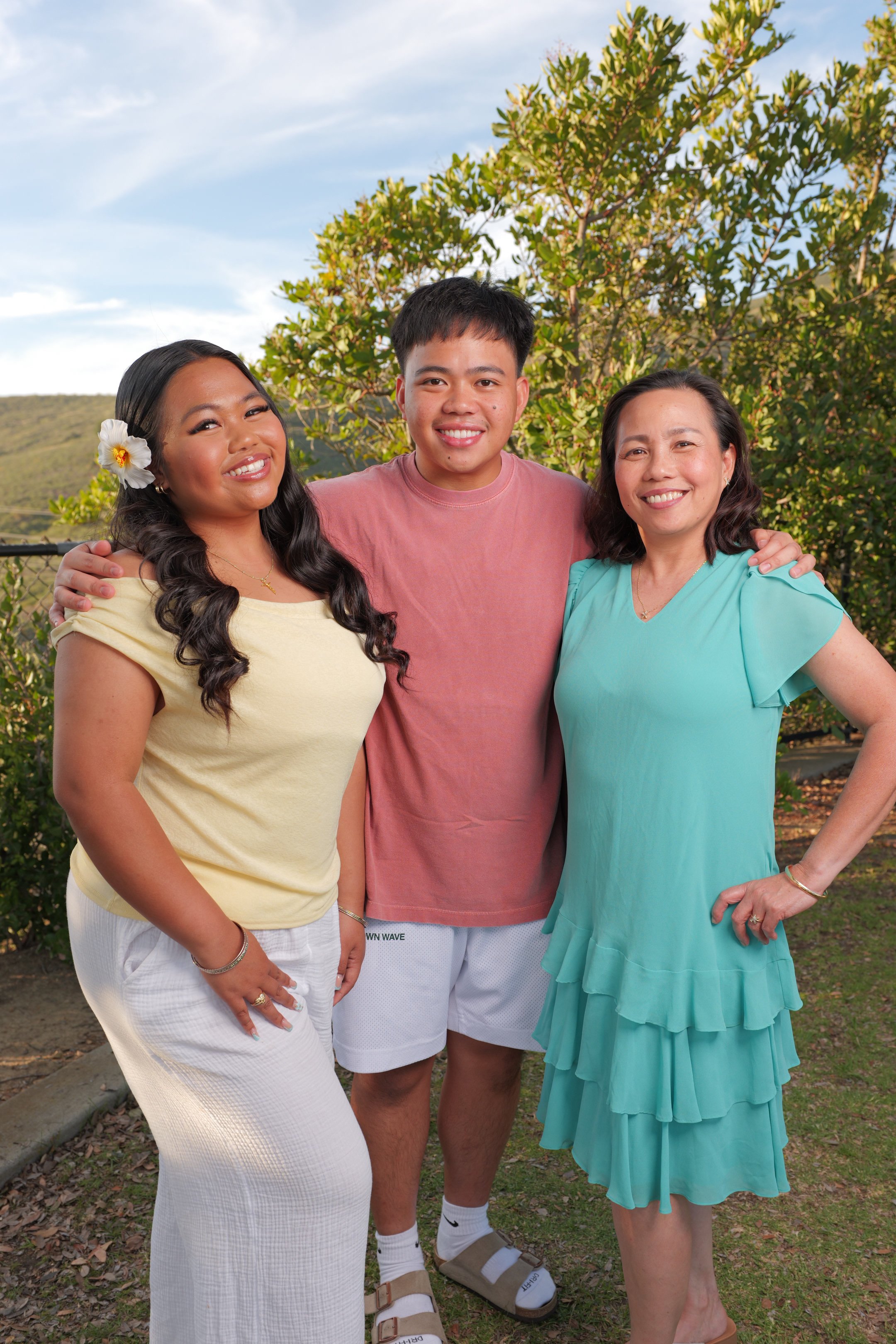 Three people, two women and one young man, standing outdoors with greenery and a blue sky in the background. They are smiling and have their arms around each other.