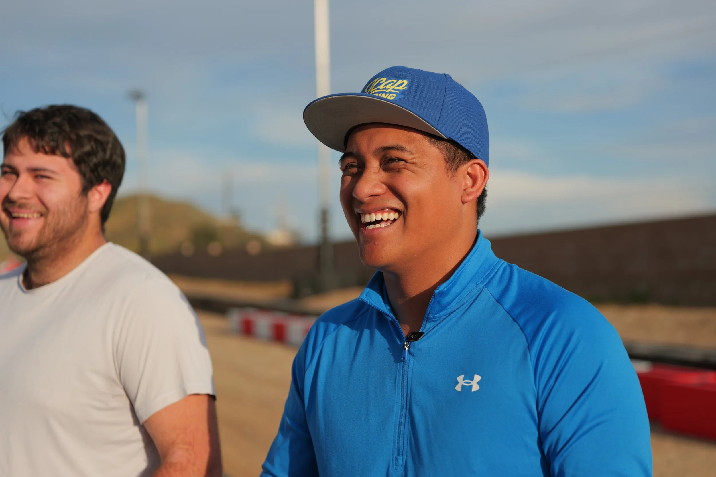 Two men smiling outdoors, one wearing a blue Under Armour zip-up jacket and a blue baseball cap with yellow writing, the other wearing a light-colored T-shirt, with a race track or outdoor setting in the background.