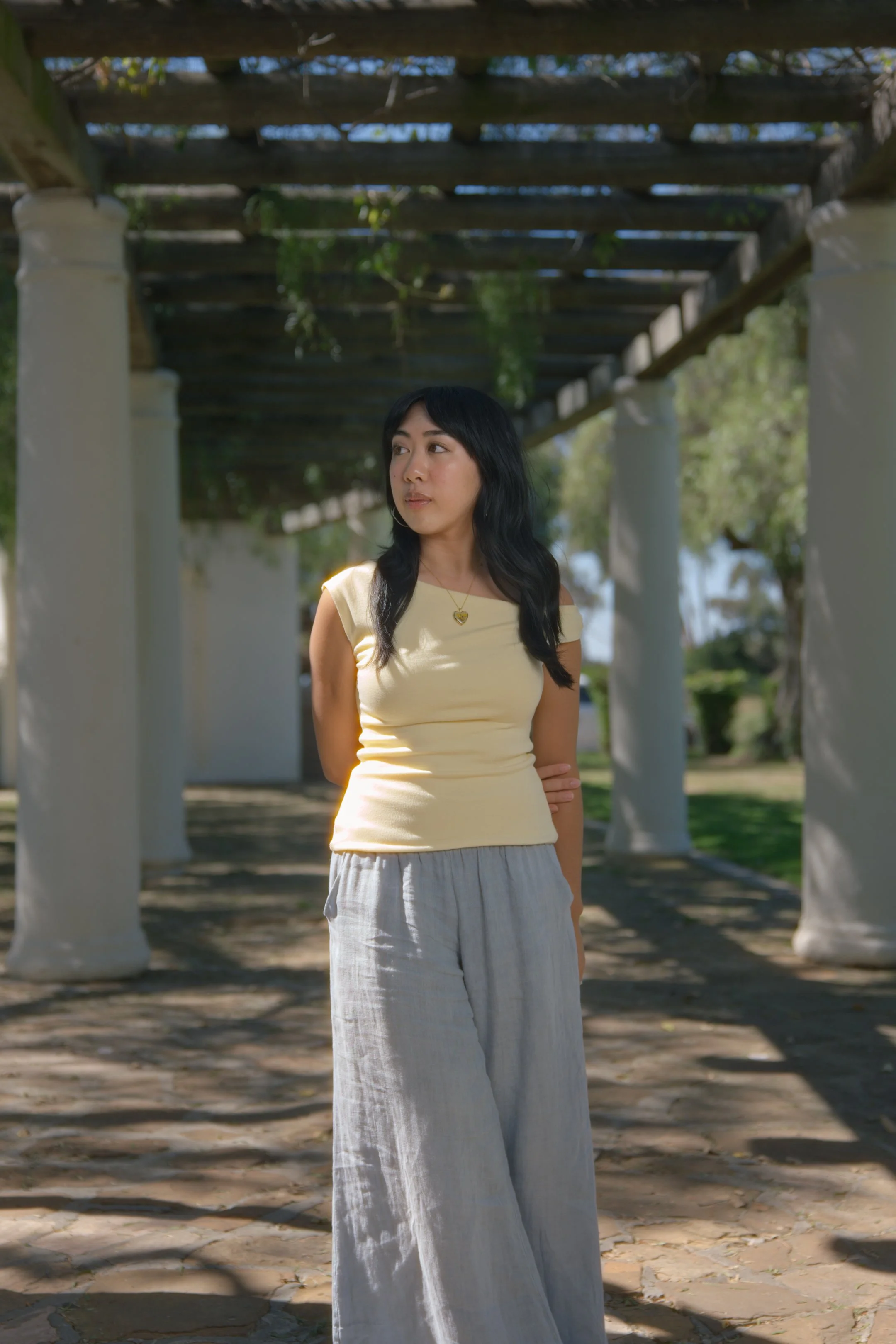 A woman with black hair wearing a yellow sleeveless top and gray wide-leg pants standing outdoors under a wooden pergola with white columns and green foliage, looking to her left.