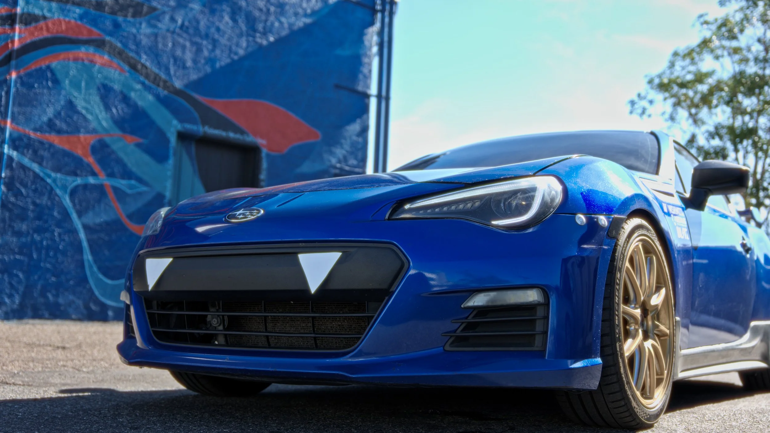 Blue Subaru sports car with gold wheels parked outdoors near a blue wall featuring a colorful mural, under a partly cloudy sky.