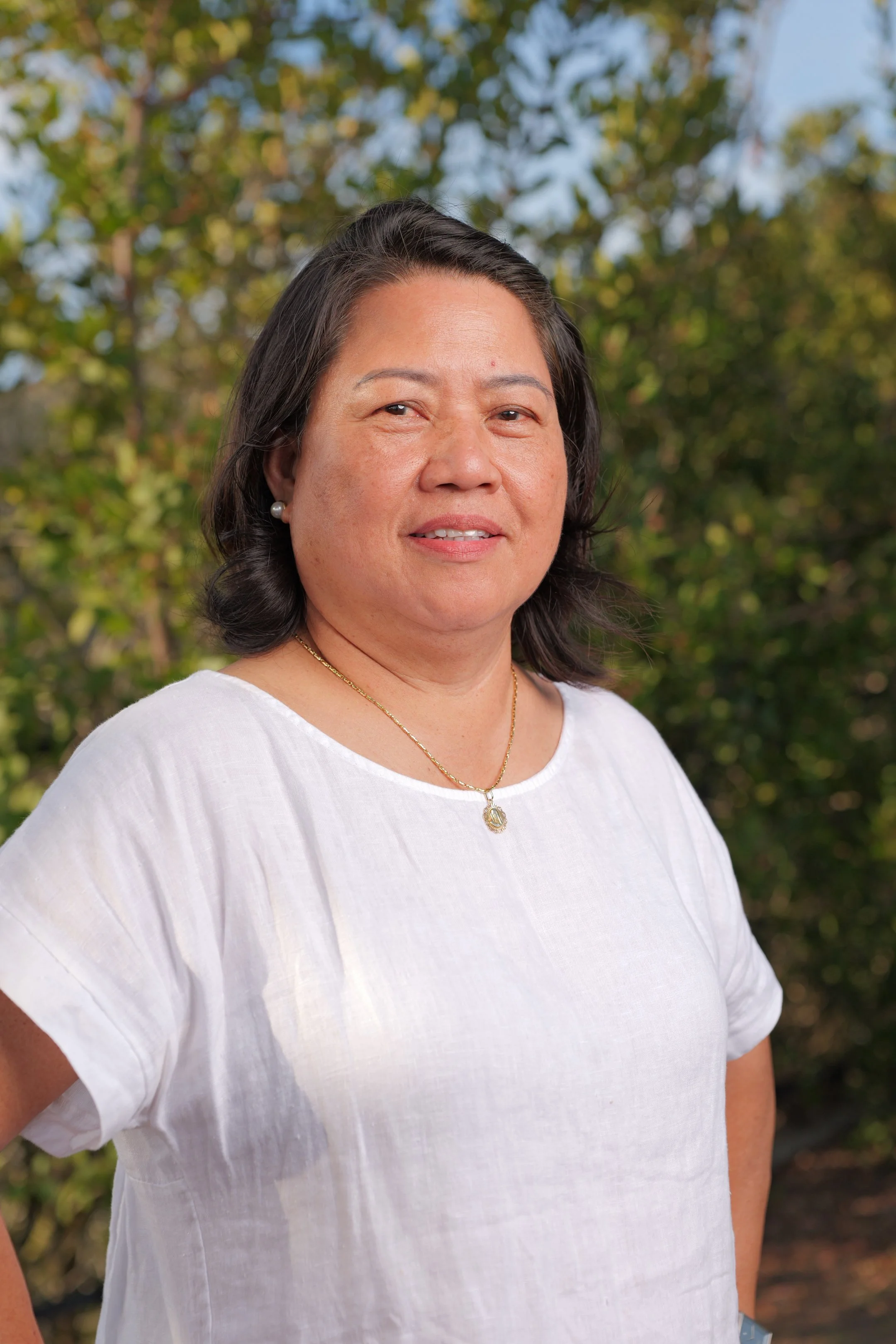 A woman in a white shirt posing outdoors with trees in the background.