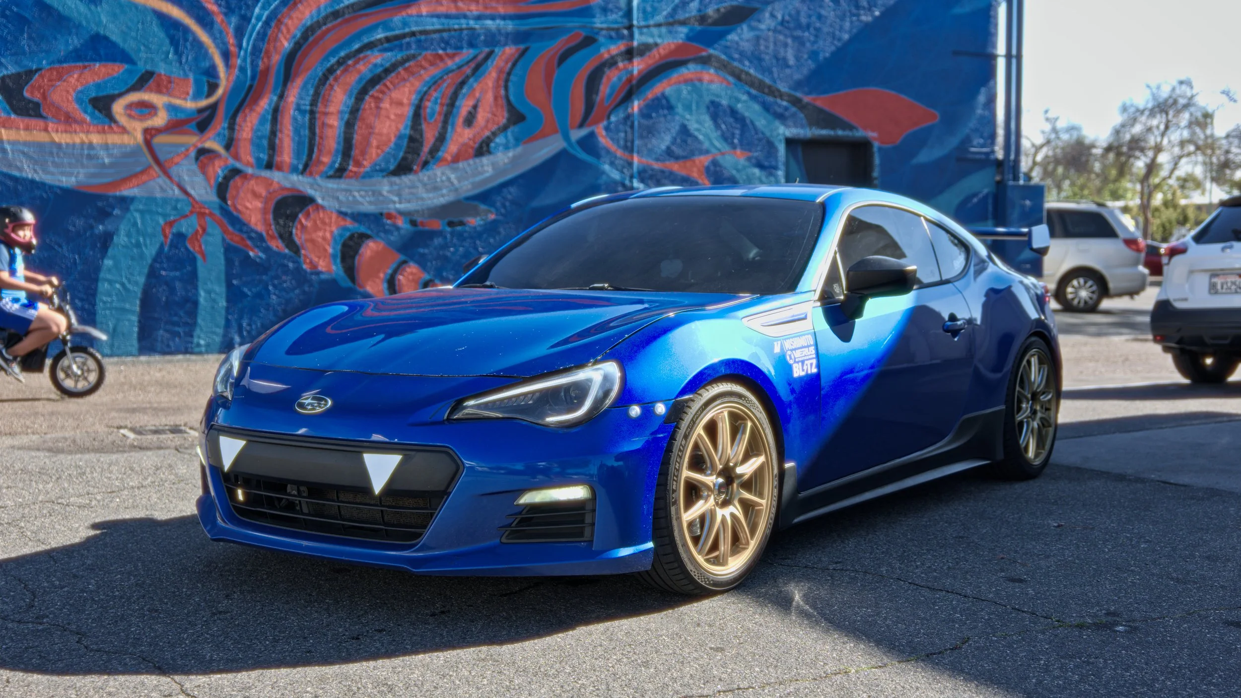 A blue sports car with gold wheels parked on the street, with a colorful mural of a fish in the background and a small child riding a bicycle nearby.