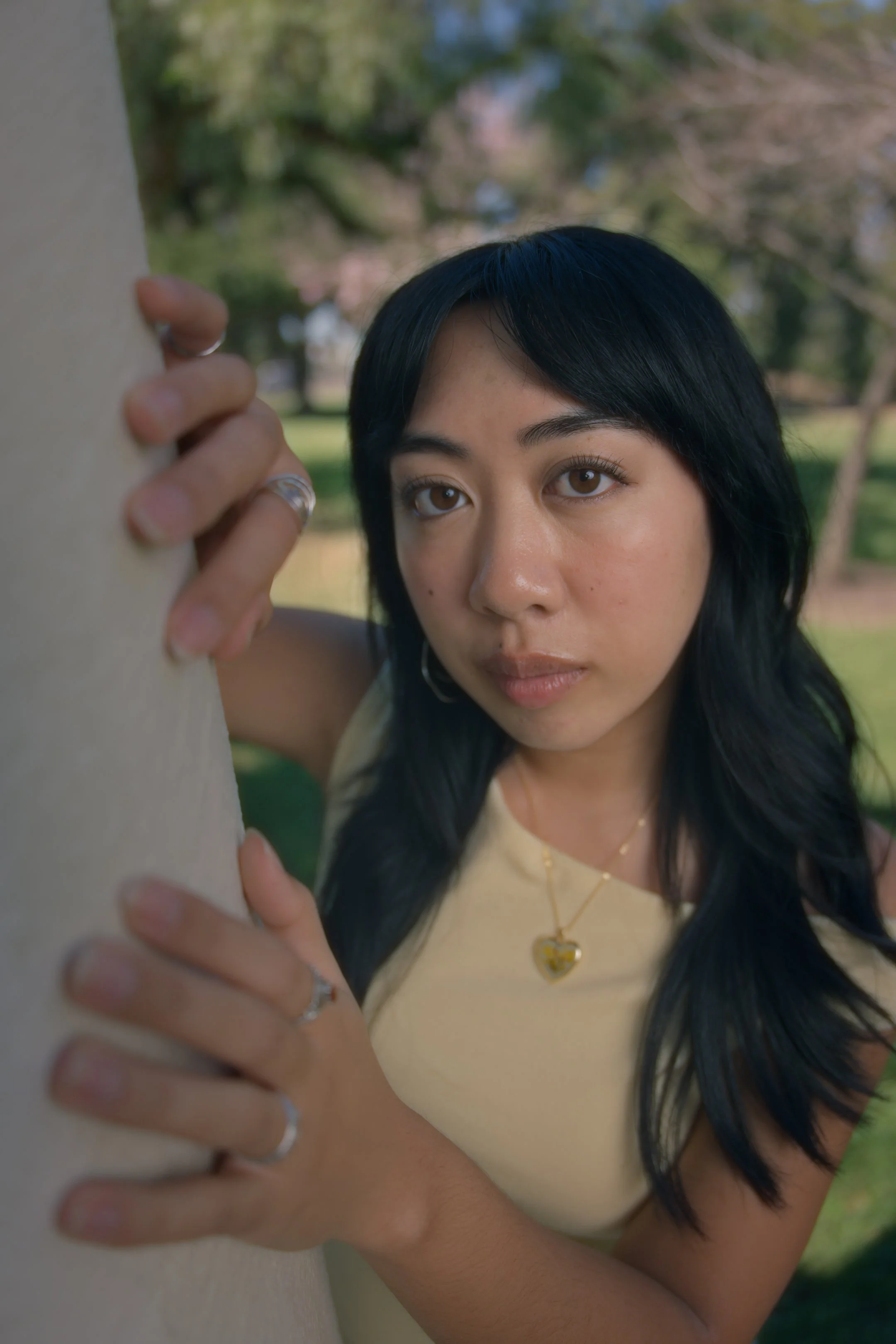 A woman with black hair and brown eyes peeks from behind a tree, looking into the distance, outdoors in a park with trees and grass.