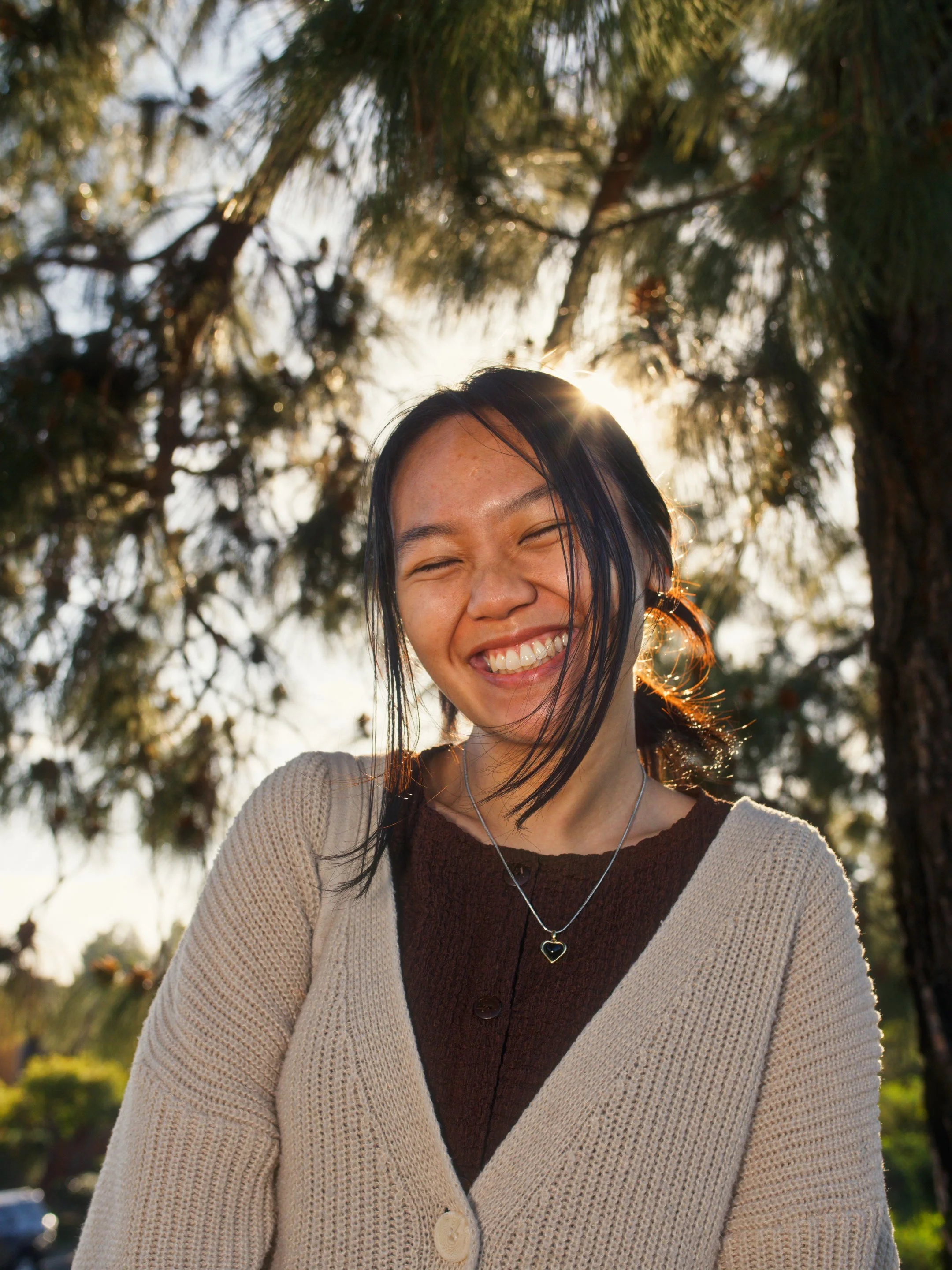 A woman with black hair smiling outdoors with sunlight filtering through pine trees behind her.