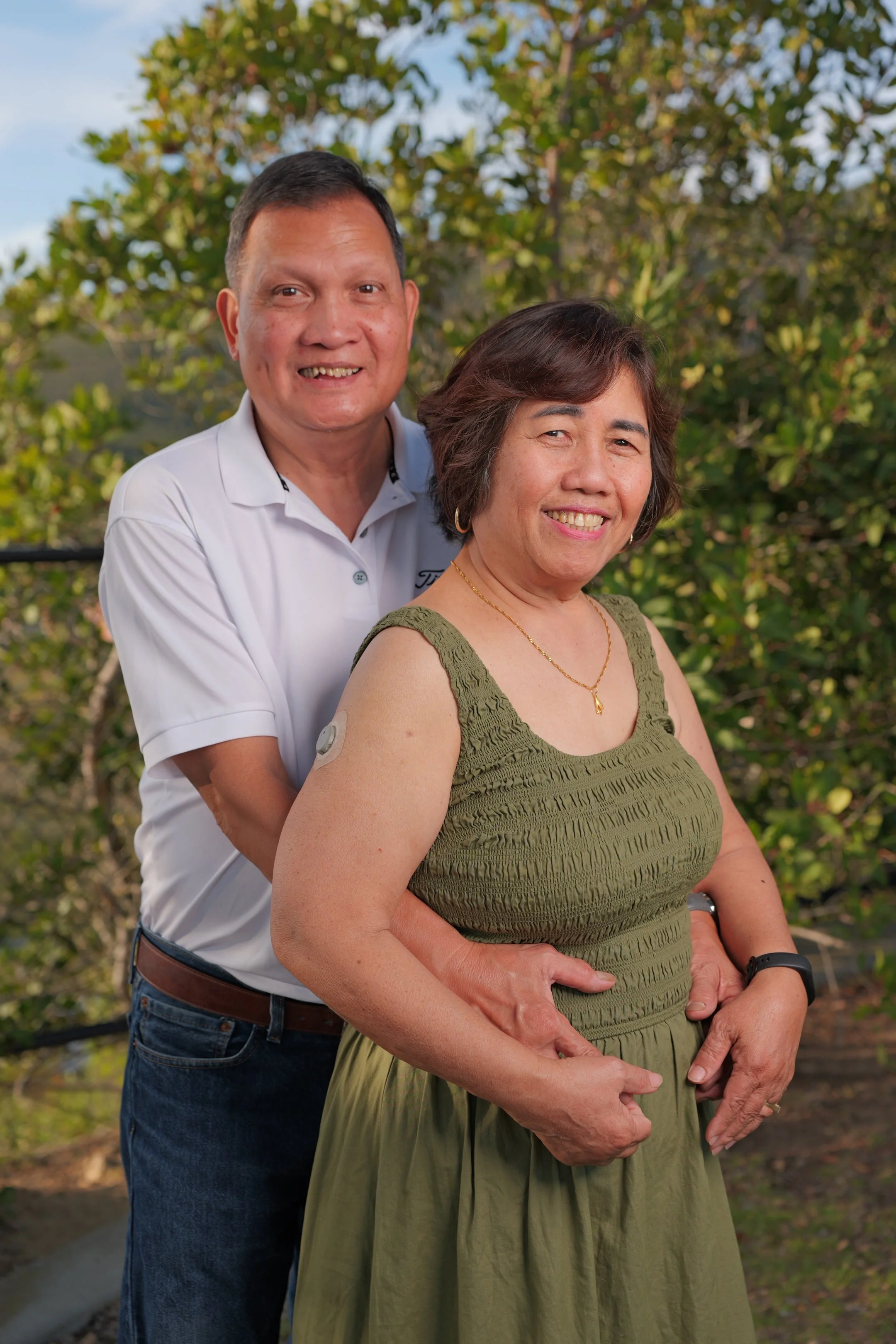 A smiling man and woman in an embrace outdoors, with green foliage and trees in the background.