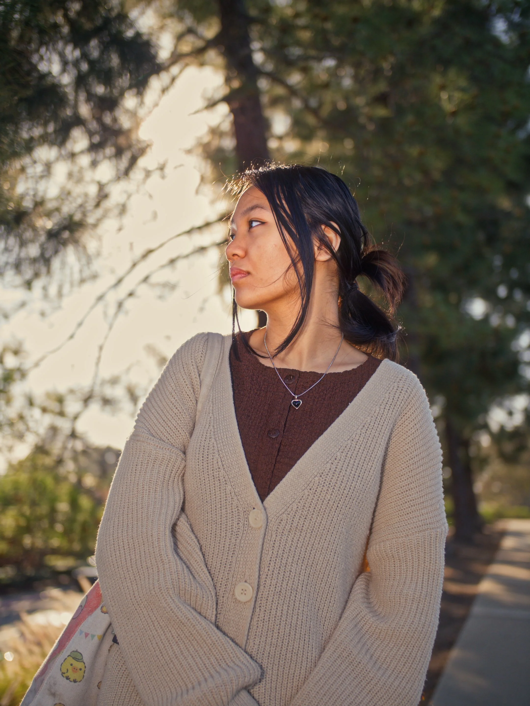 A woman with dark hair in a ponytail is standing outdoors in front of trees, looking to the side. She is wearing a beige cardigan over a brown top and has a necklace with a heart-shaped pendant.