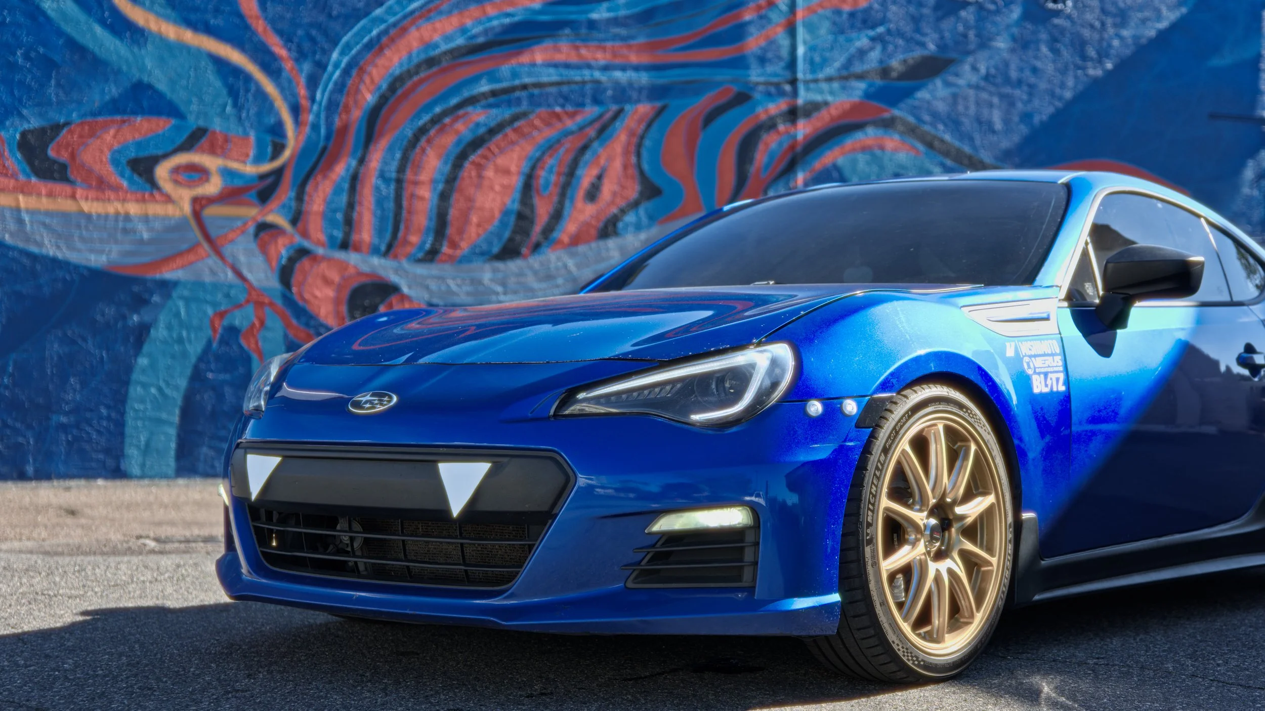 A blue Subaru sports car with gold wheels parked in front of a colorful mural featuring a dragon.