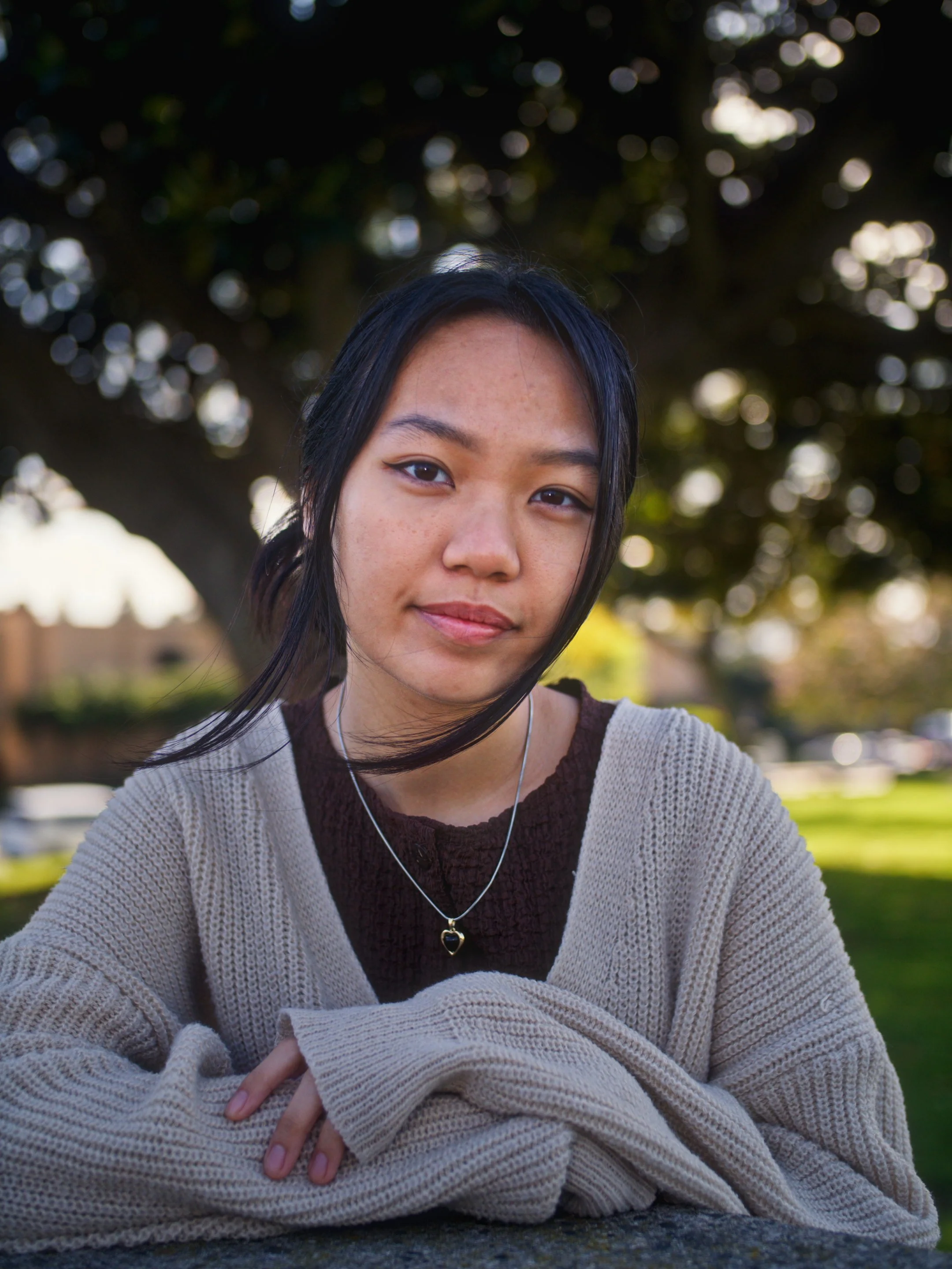 A young woman with black hair, wearing a beige cardigan and a necklace with a heart-shaped pendant, sitting outdoors near a park bench with a large tree and grass in the background.
