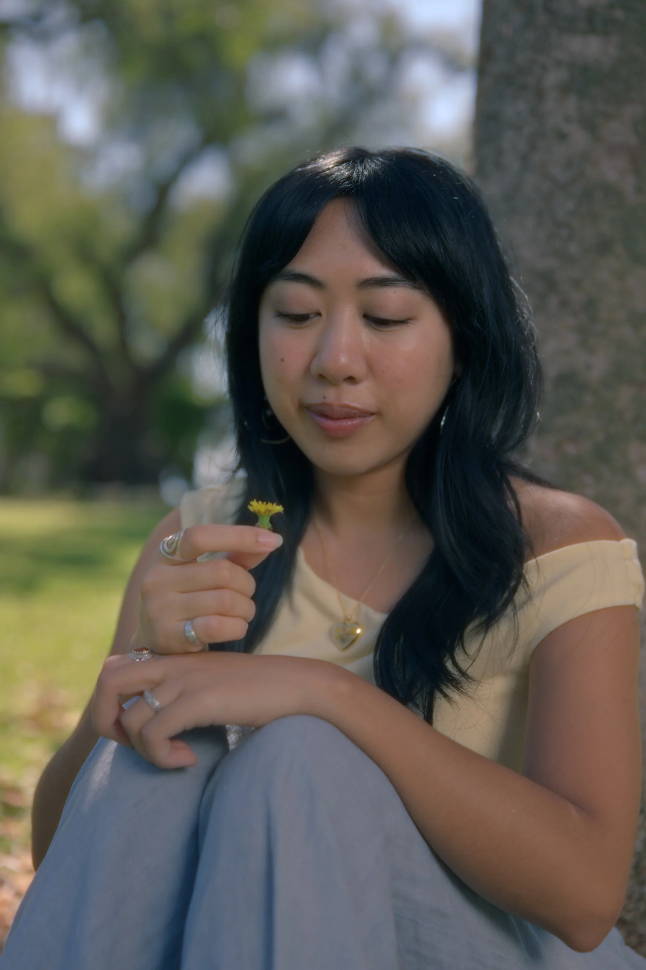 A woman with black hair and multiple rings on her fingers, sitting outdoors next to a tree, holding a small yellow flower, and looking at it gently.