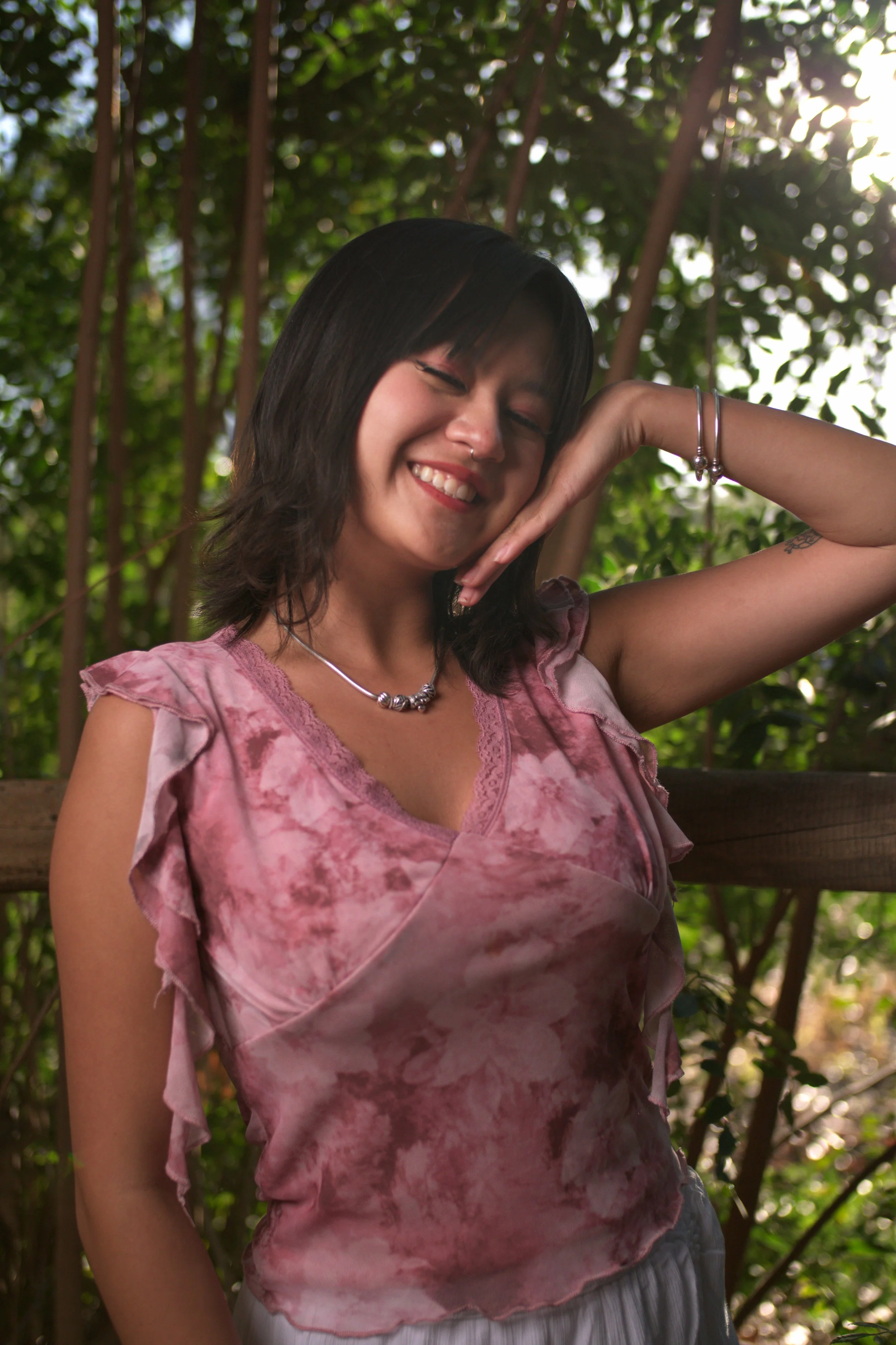 Young woman in pink tie-dye blouse smiling and posing with her hand resting on her face outdoors with trees and sunlight in the background.