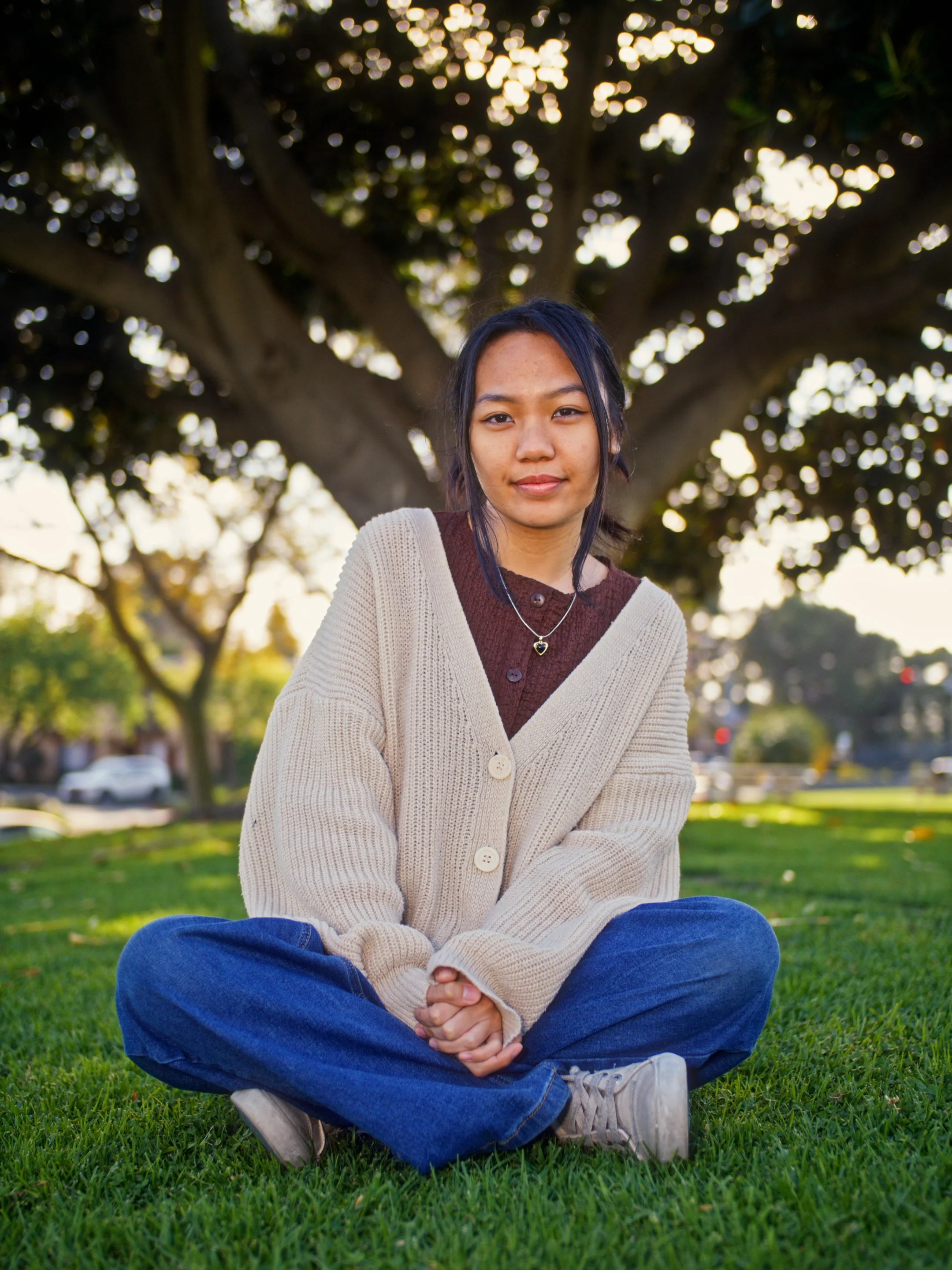 Young woman sitting cross-legged on grass in a park with a large tree behind her, wearing a beige cardigan and blue jeans, during late afternoon or early evening.