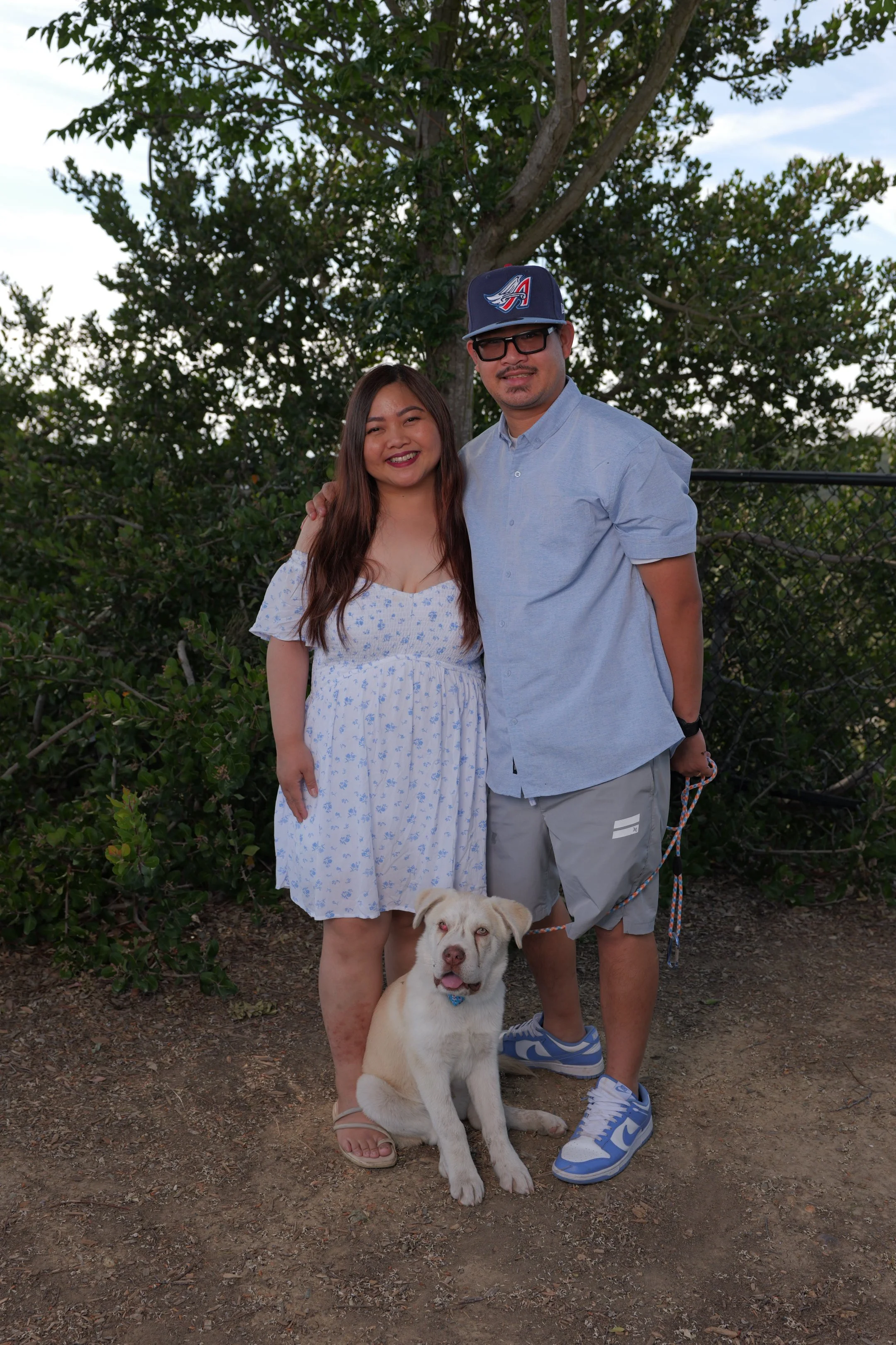 A woman and a man standing outdoors with a dog in front, smiling, with trees and a chain-link fence in the background during daytime.