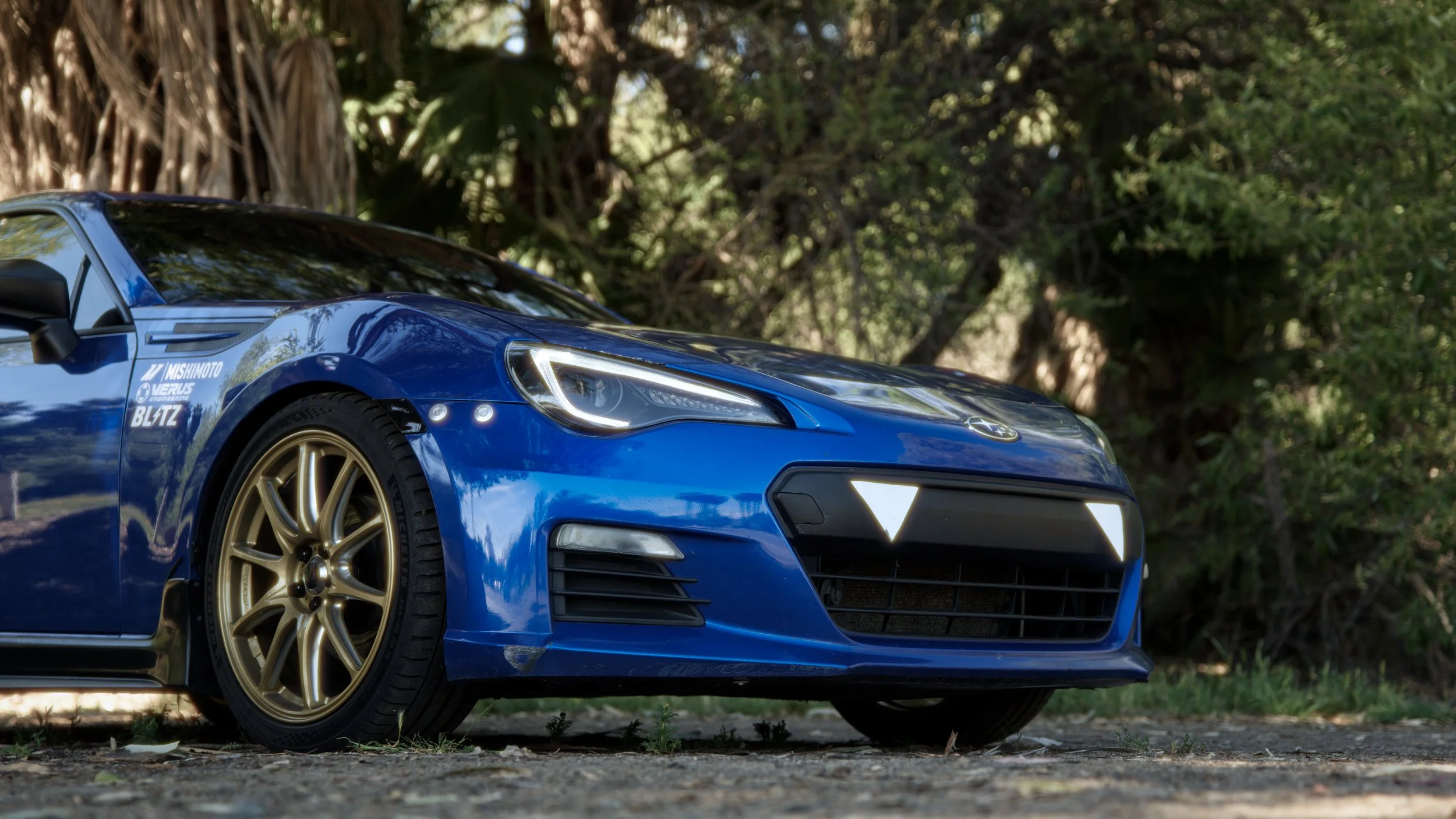 Close-up of a blue sports car with gold wheels parked on a dirt surface with greenery in the background.