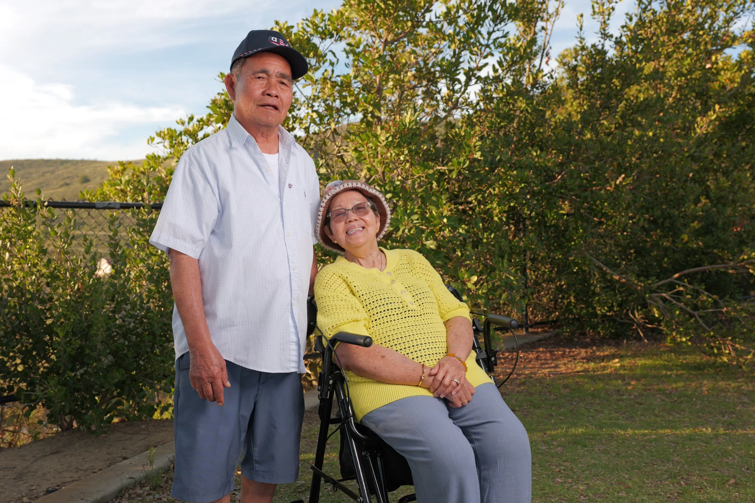 An elderly woman with glasses sitting in a wheelchair, wearing a yellow sweater and a hat, smiling. Beside her, an elderly man wearing a white shirt, gray shorts, and a cap, standing outdoors with green bushes and hills in the background.