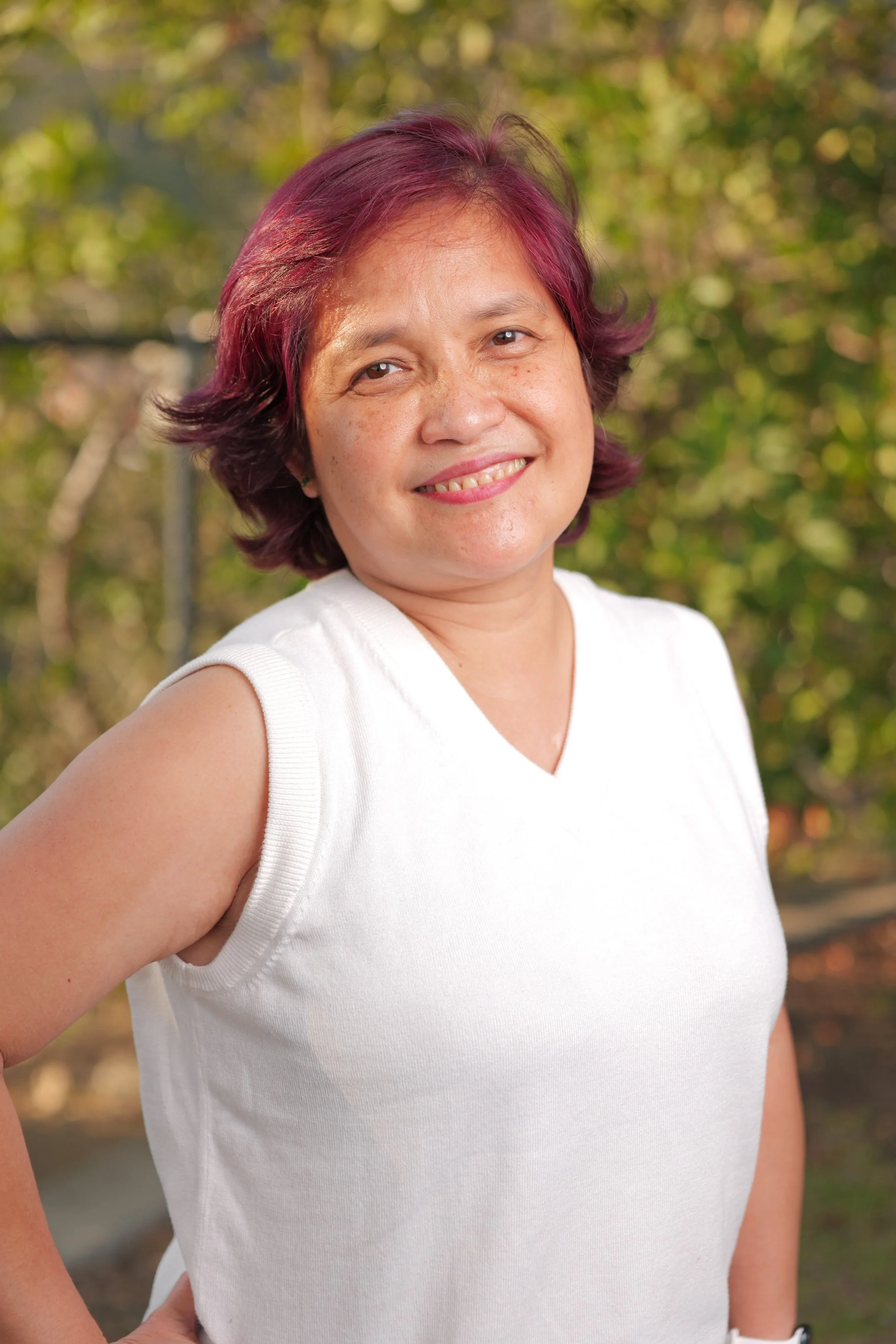 A woman with short, dark pink hair smiling outdoors with greenery in the background, wearing a white sleeveless top.