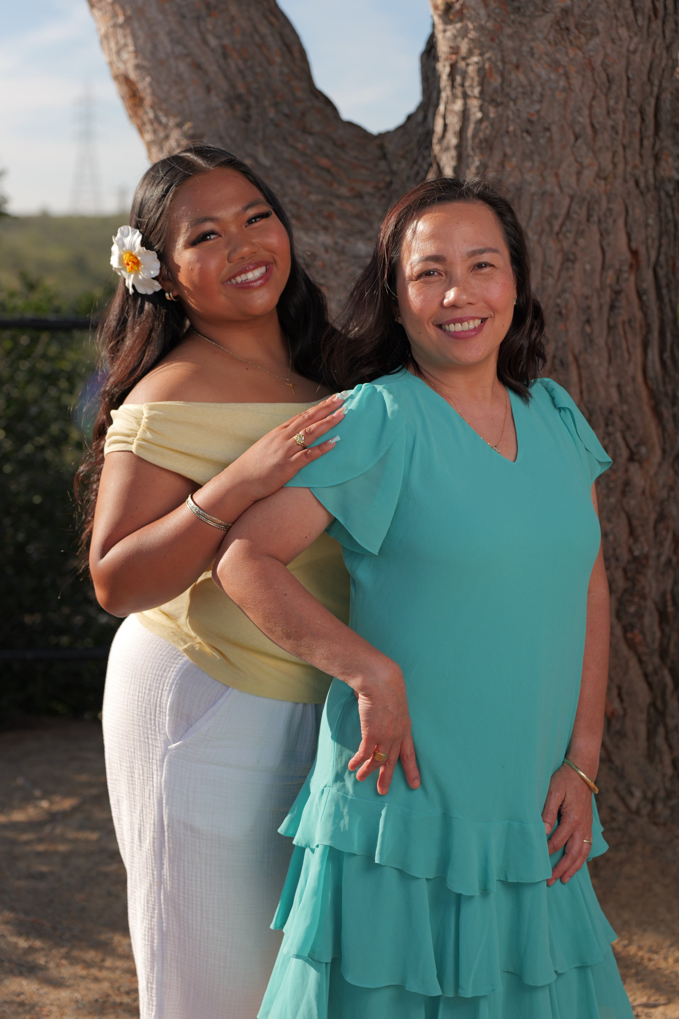 Two women smiling outdoors in front of a tree, one with long dark hair and the other with shoulder-length dark hair, wearing colorful dresses.