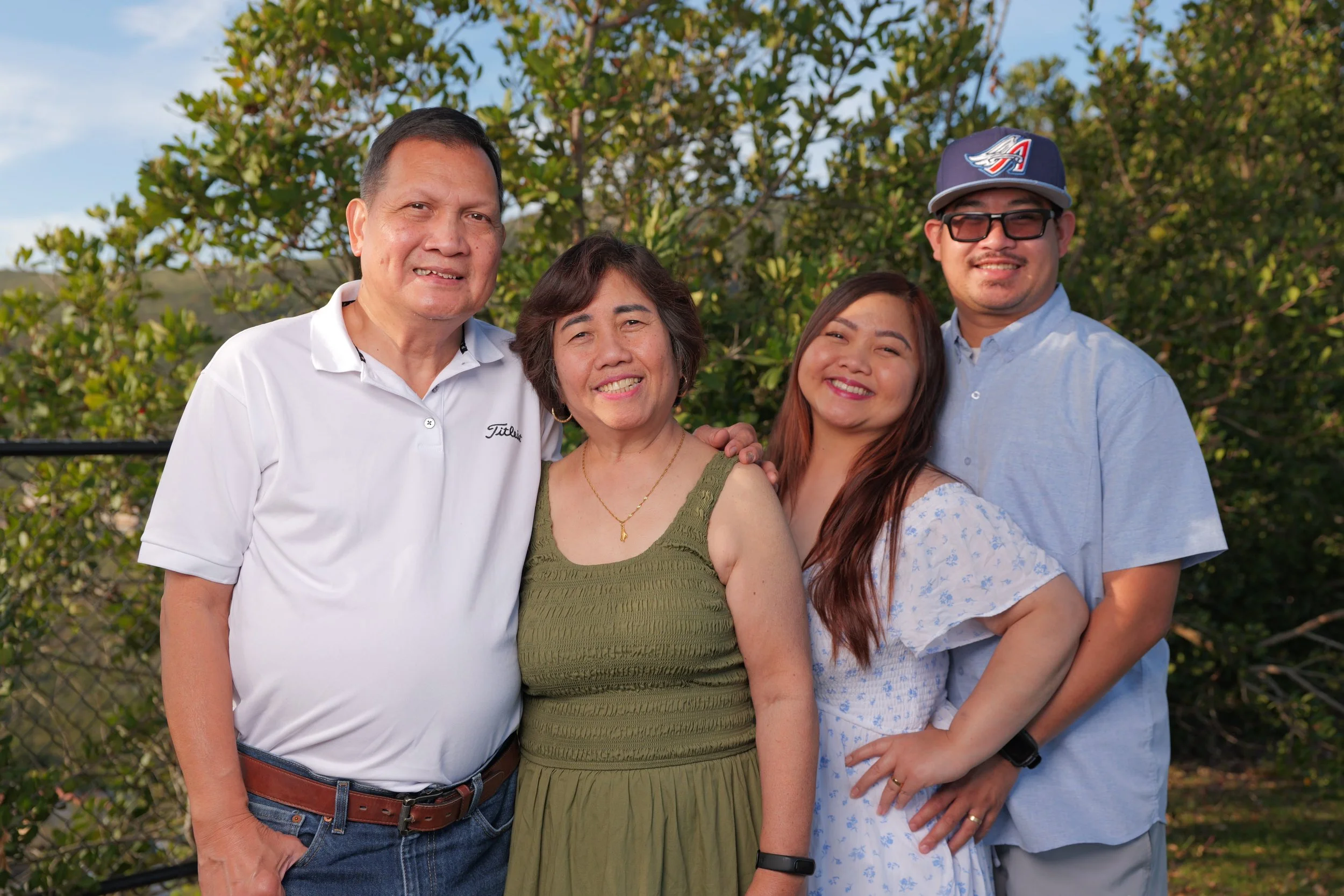 Family of five standing outdoors near a black fence with green trees and blue sky in the background, smiling at the camera.