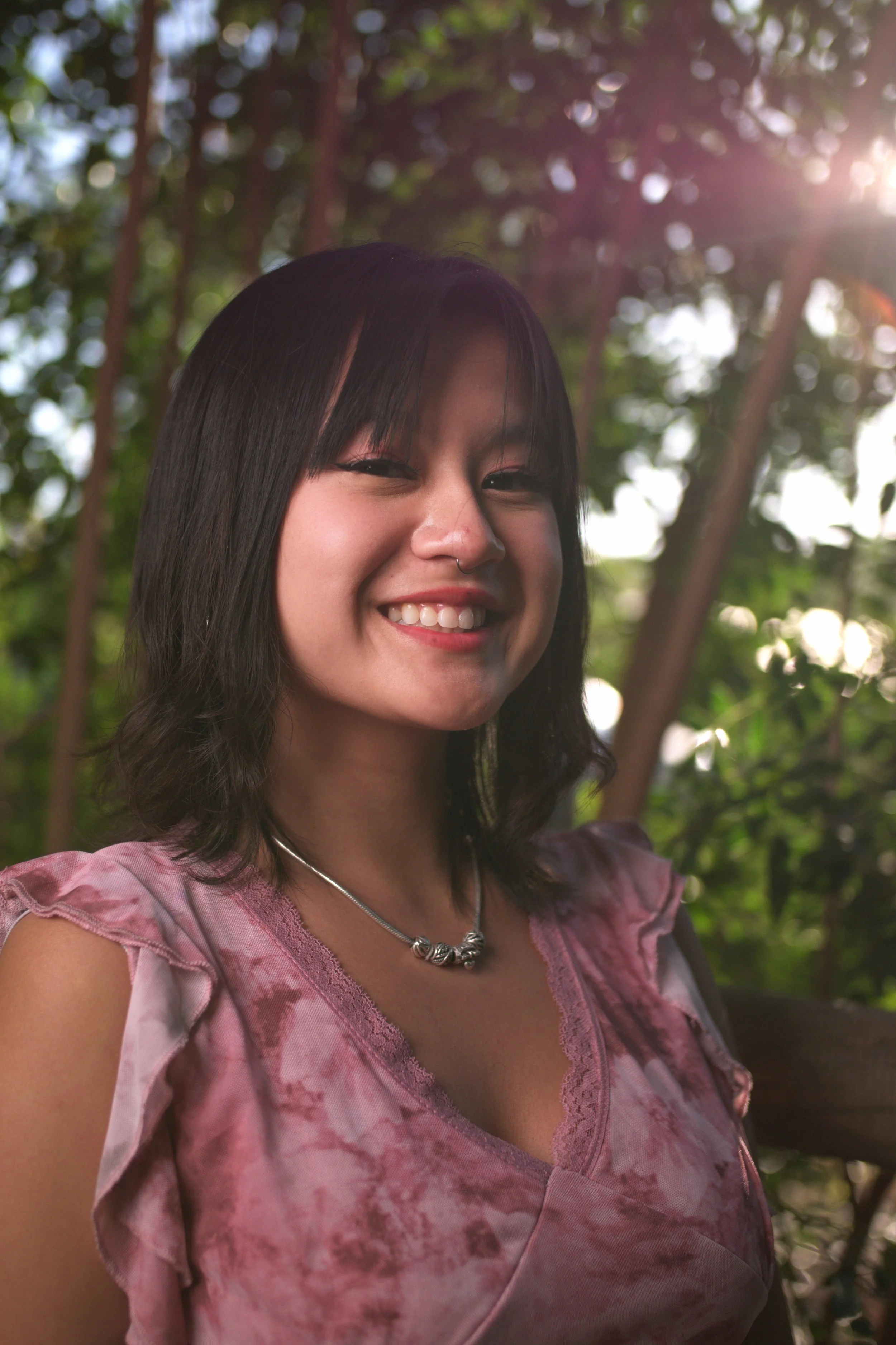 A smiling young woman with black hair and a nose ring, wearing a pink and purple tie-dye top and a silver necklace, outdoors with green foliage and sunlight in the background.