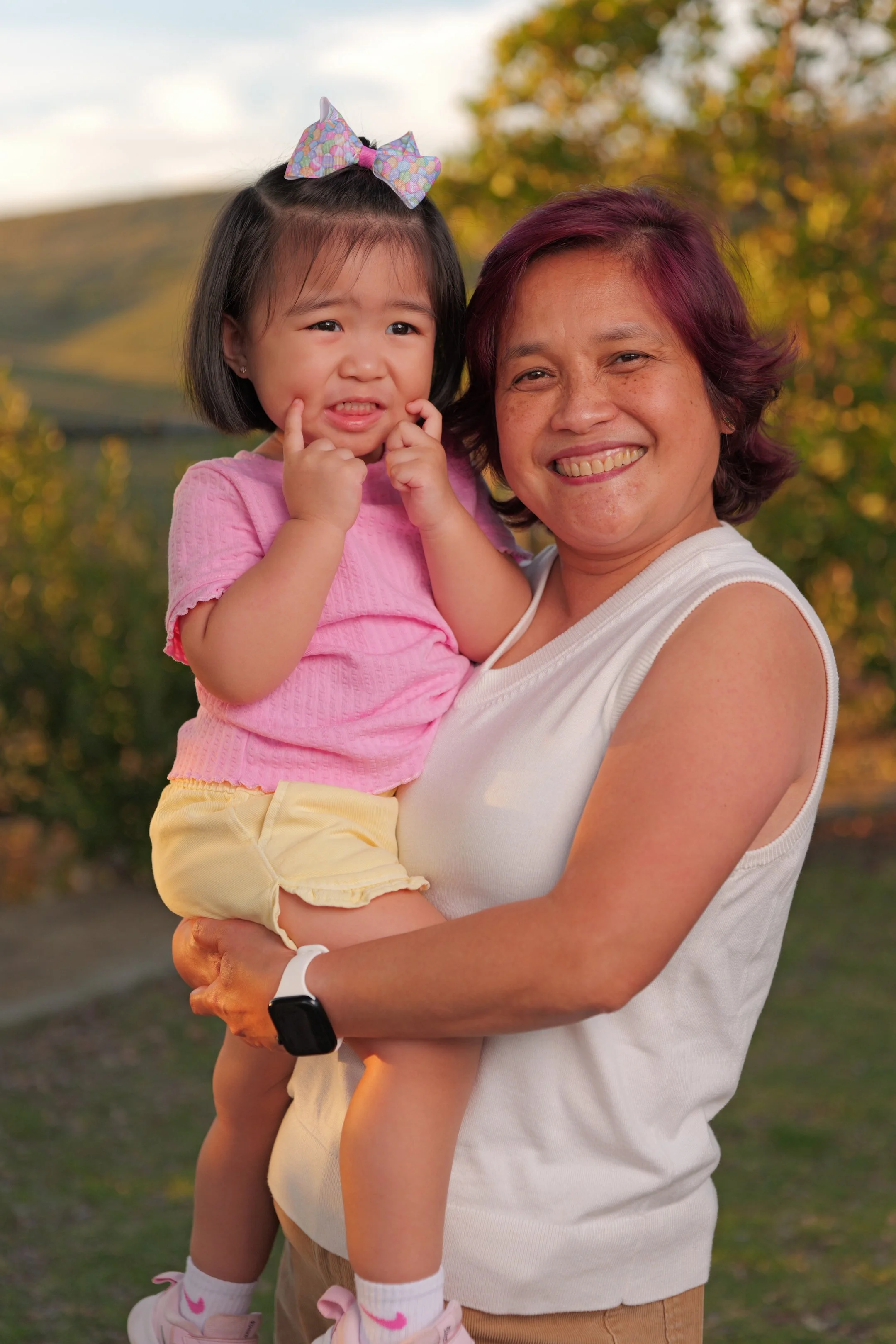 A woman with short purple hair and a white sleeveless top smiling while holding a young girl in pink clothing with a colorful bow in her hair outdoors during fall.