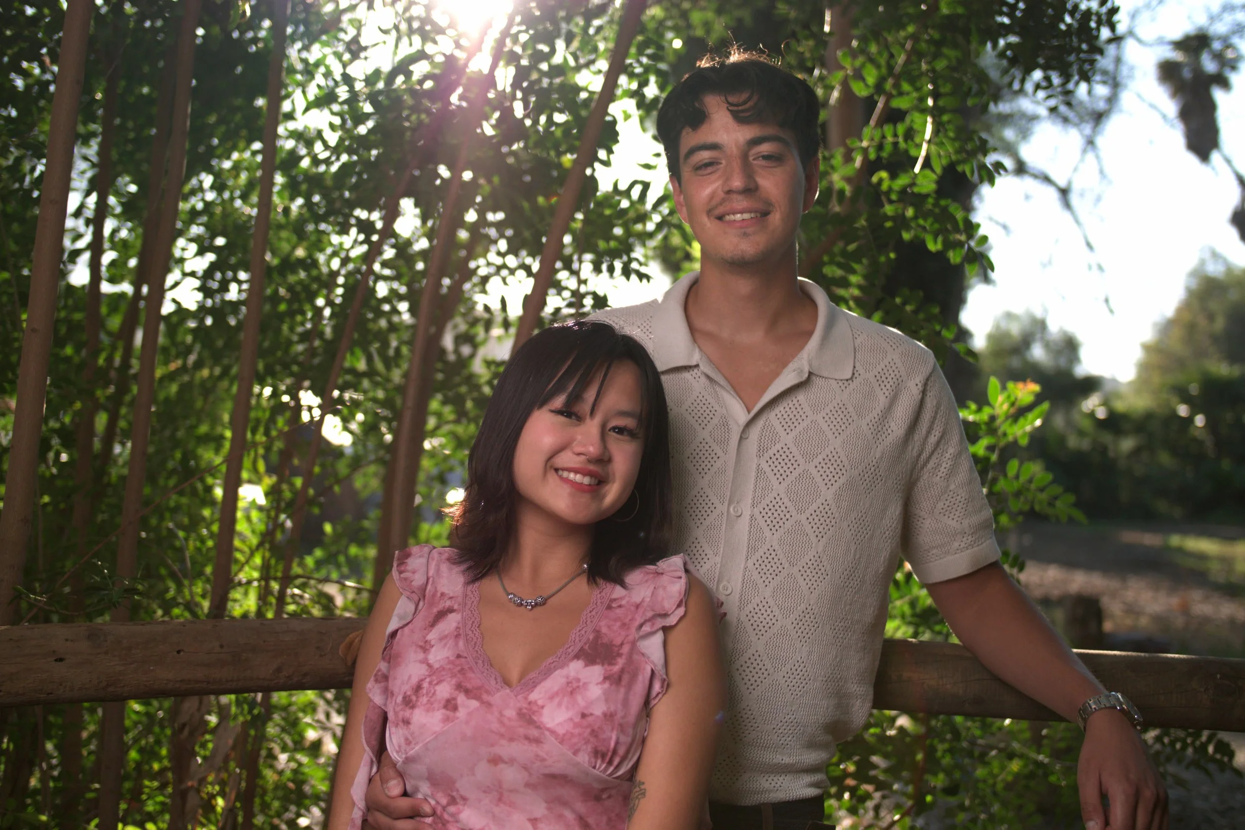 A smiling young woman and man standing together outdoors in front of green foliage, with sunlight filtering through the trees behind them.