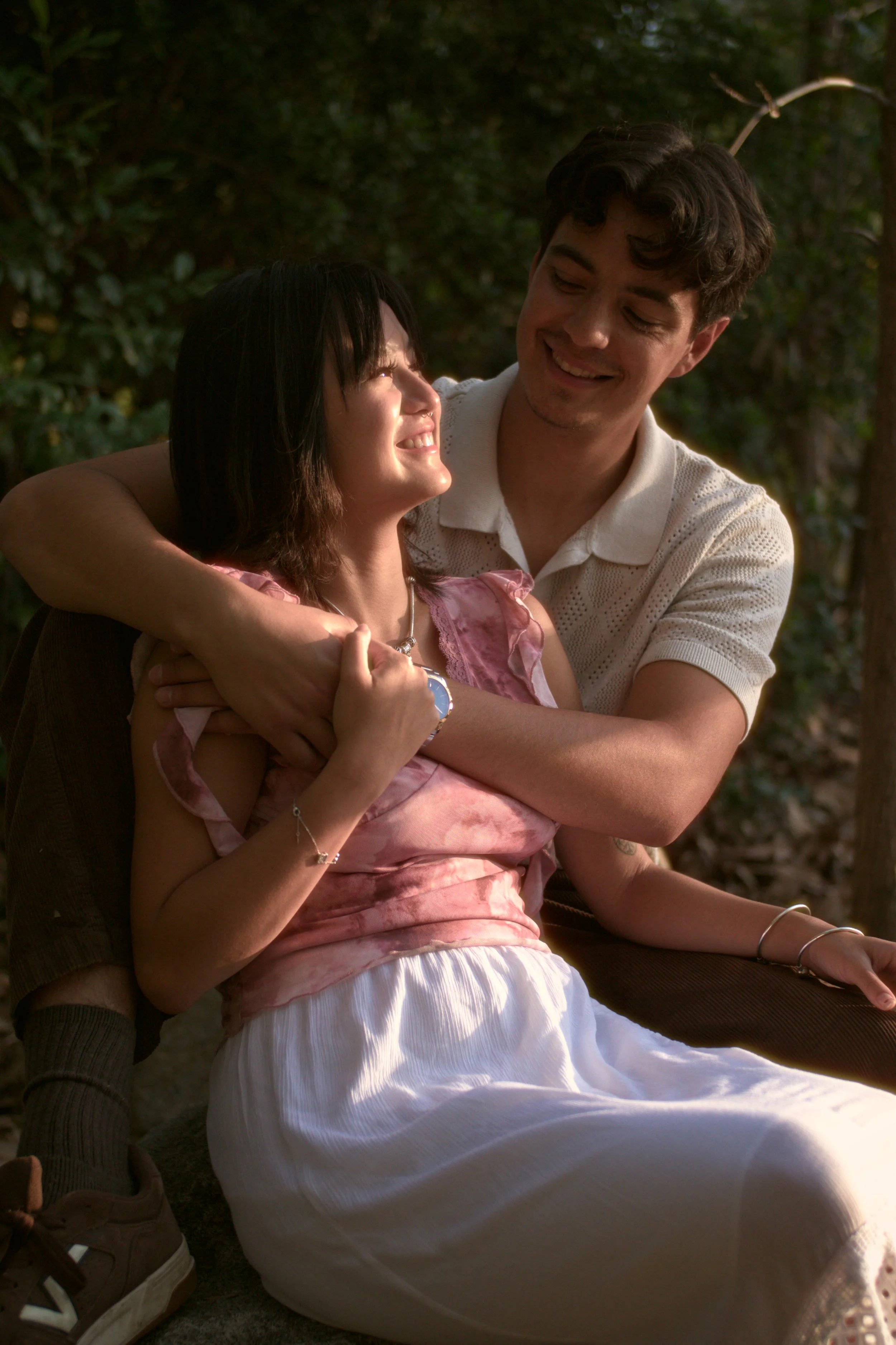 A young couple sitting outdoors, embracing and smiling at each other, surrounded by greenery.