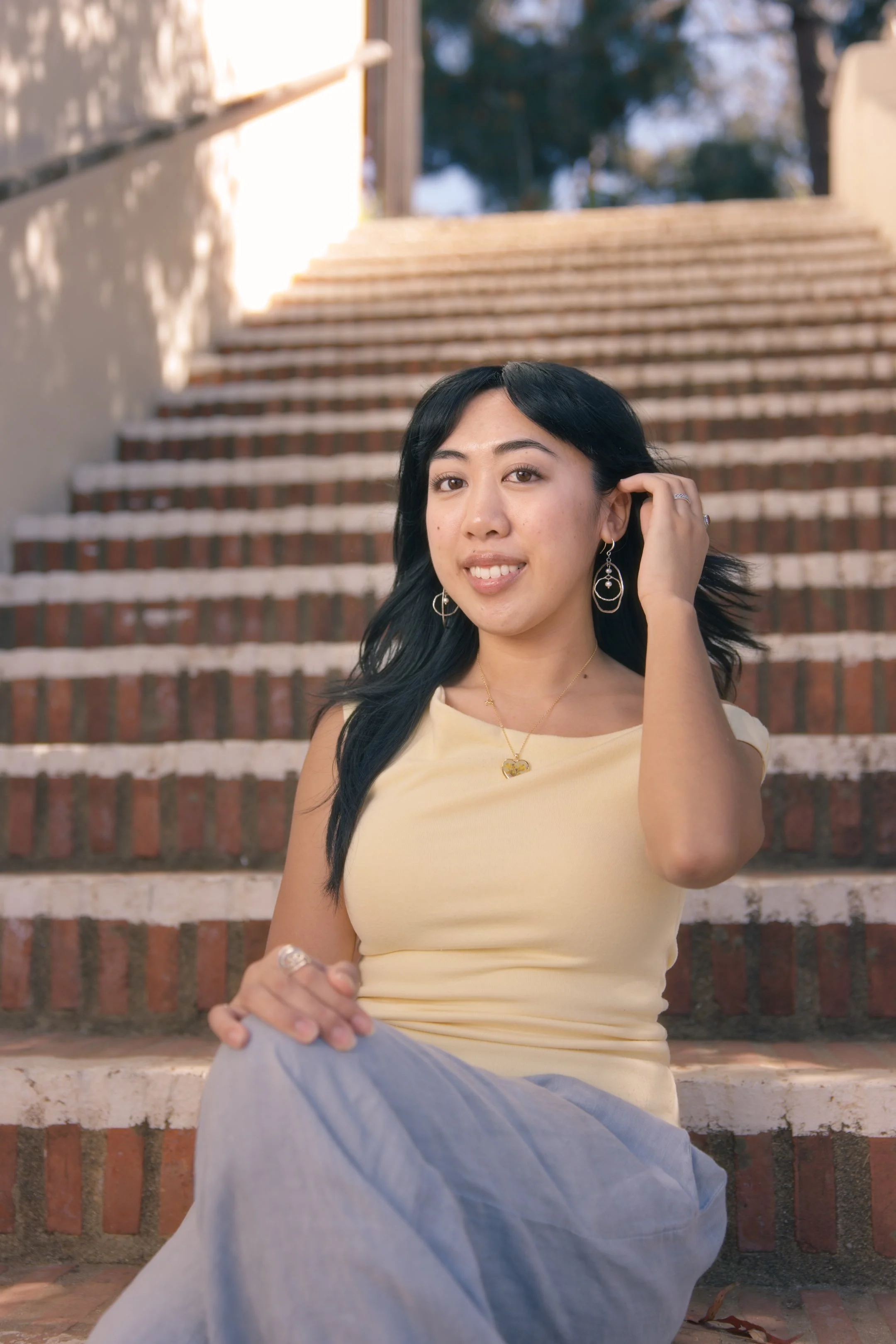 A young woman sitting on brick steps outdoors, wearing a yellow top and gray pants, touching her black hair with her right hand, and smiling at the camera.