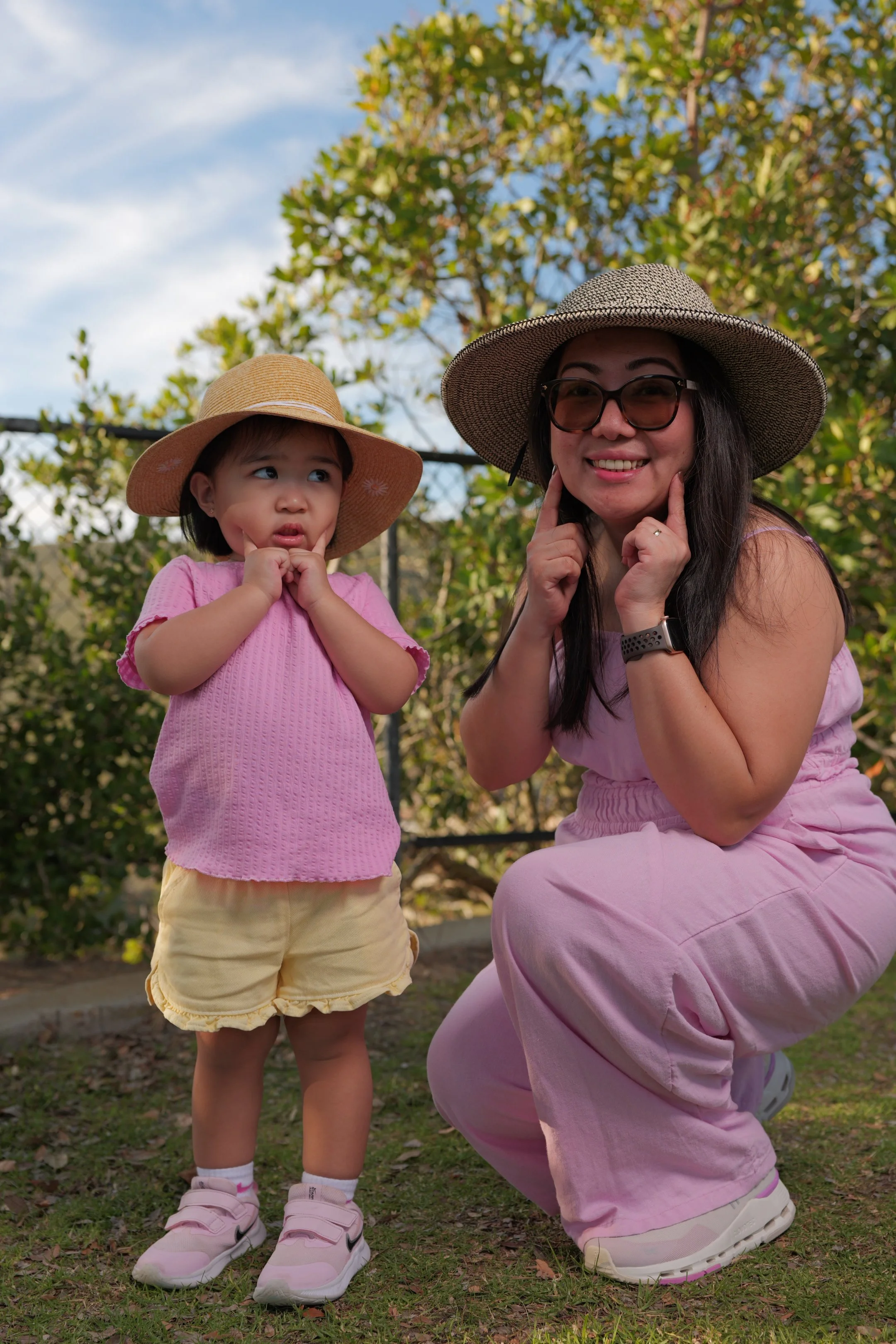 A woman and a young girl outdoors wearing hats, with greenery and sky in the background. The woman is crouching, smiling, and touching her face, while the girl looks confused or curious, touching her face with both hands.