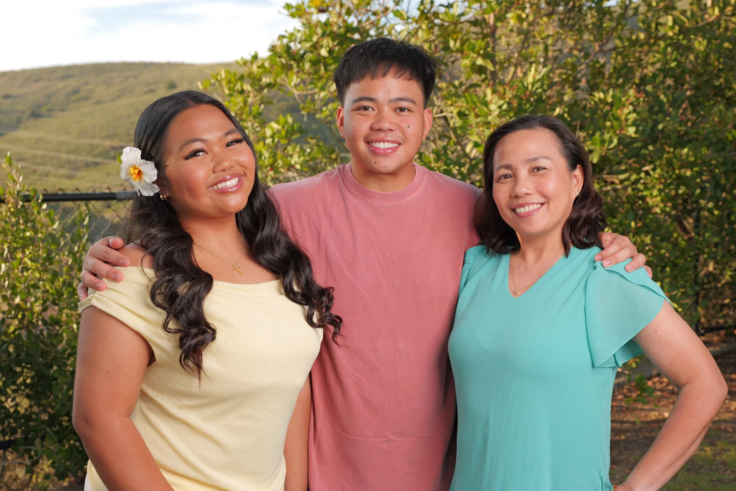 Three people smiling outdoors, standing close together with arms around each other, with trees and hills in the background.