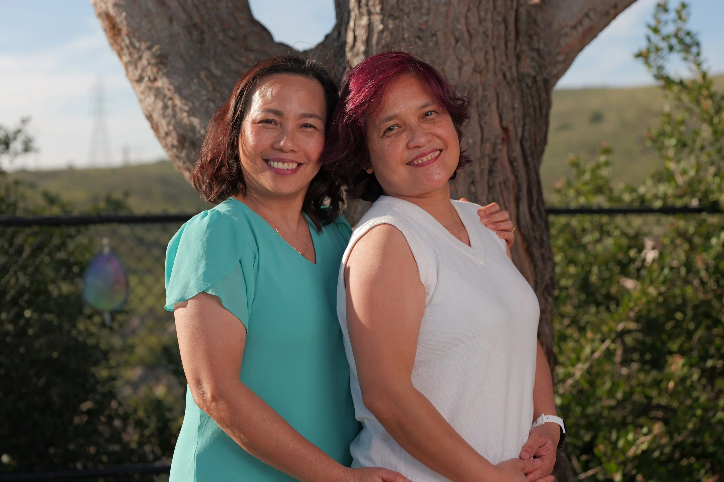 Two women standing outdoors in front of a large tree, smiling at the camera. One woman has short dark hair and is wearing a teal top, the other has short reddish hair and is wearing a white sleeveless top.