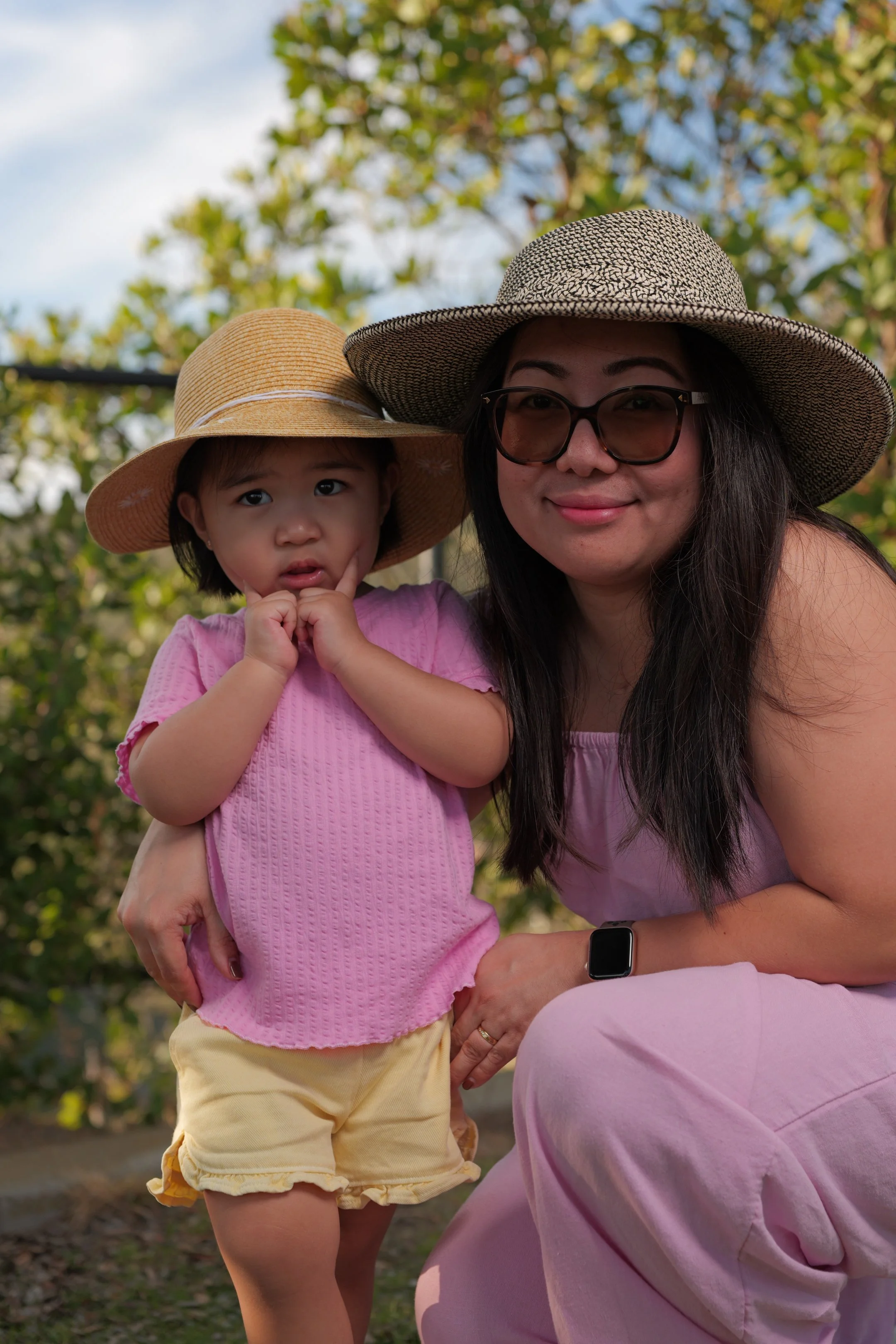 A woman and a young girl wearing wide-brimmed hats and pink clothing outdoors with trees in the background.