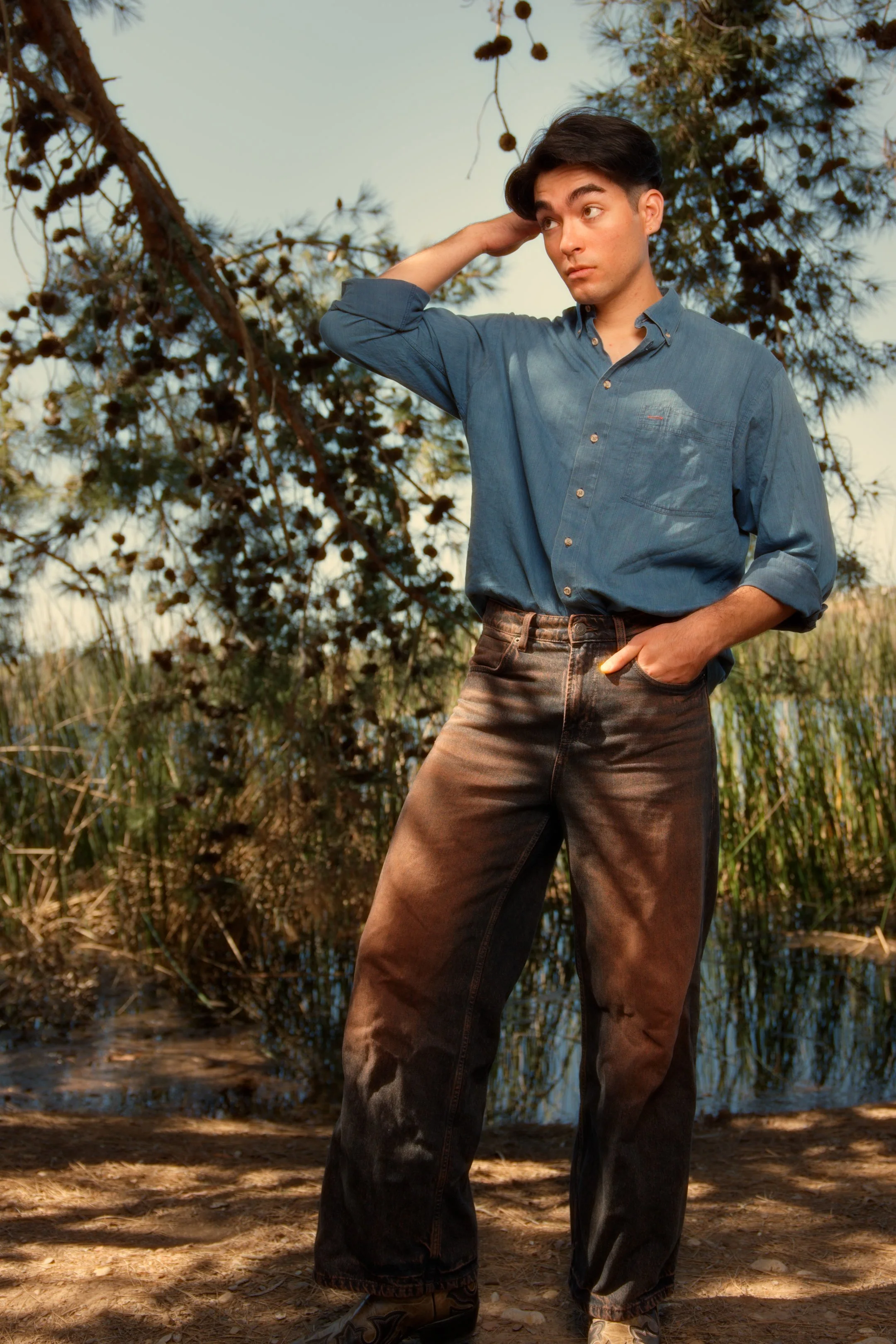 A young man standing outdoors near a body of water, with a large tree and tall grasses in the background. He is wearing a blue button-up shirt and dark jeans, with one hand in his pocket and the other hand touching his head.